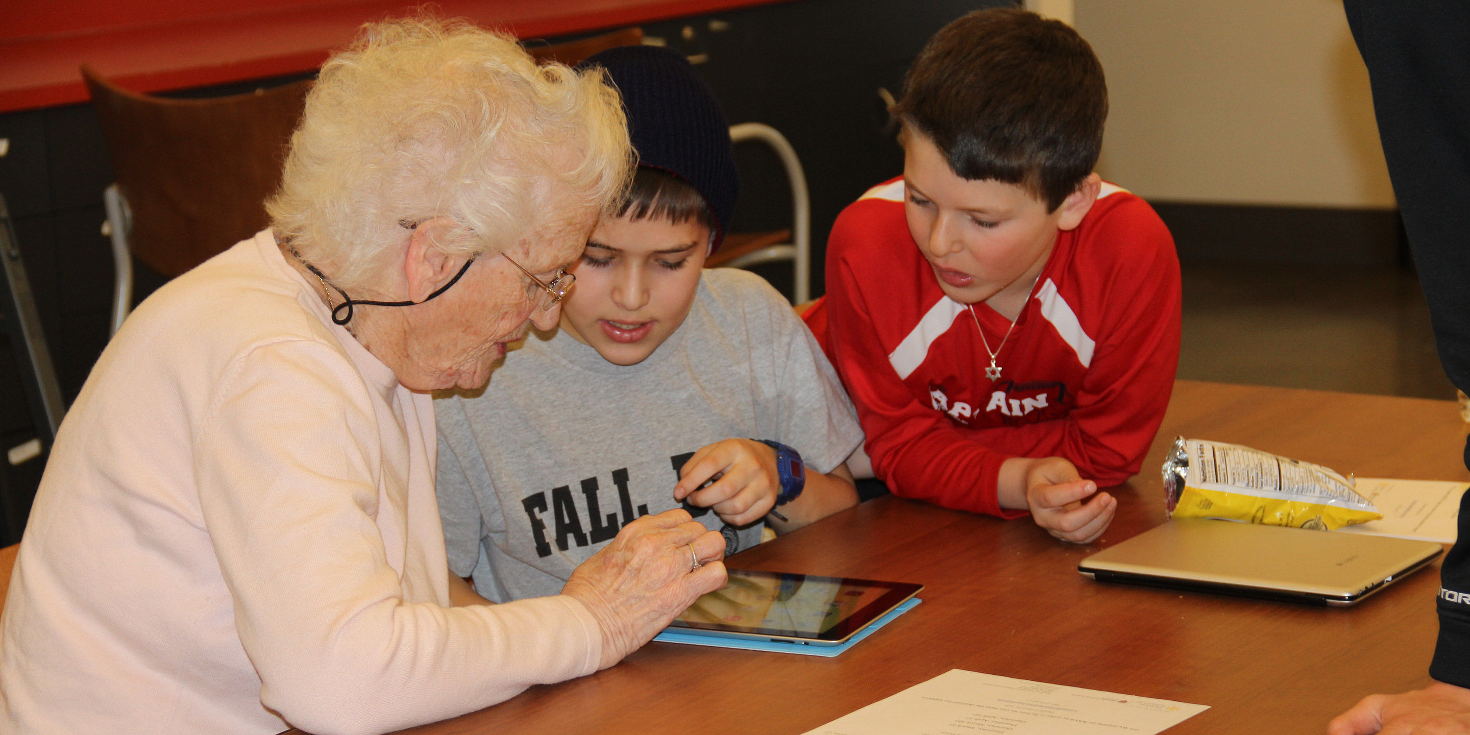 An older person and two middle school students sit at a table, looking at a tablet together.