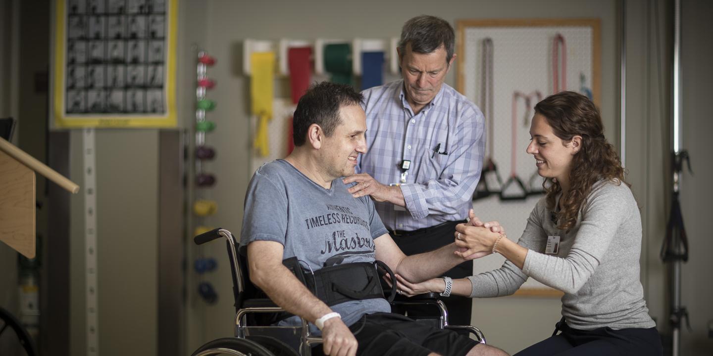 A physical therapy patient in a wheelchair in a gym at Hebrew Rehabilitation Center in Boston. A physical therapy student is crouched next to him, holding his arm and elbow. A physical therapist stands behind with his hand on the man’s shoulder.