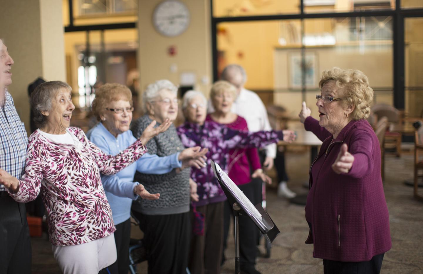 On the left is a line of older women and men who are singing. One of the women has her arms outstretch. On the right facing them is another older woman who is conducting them.