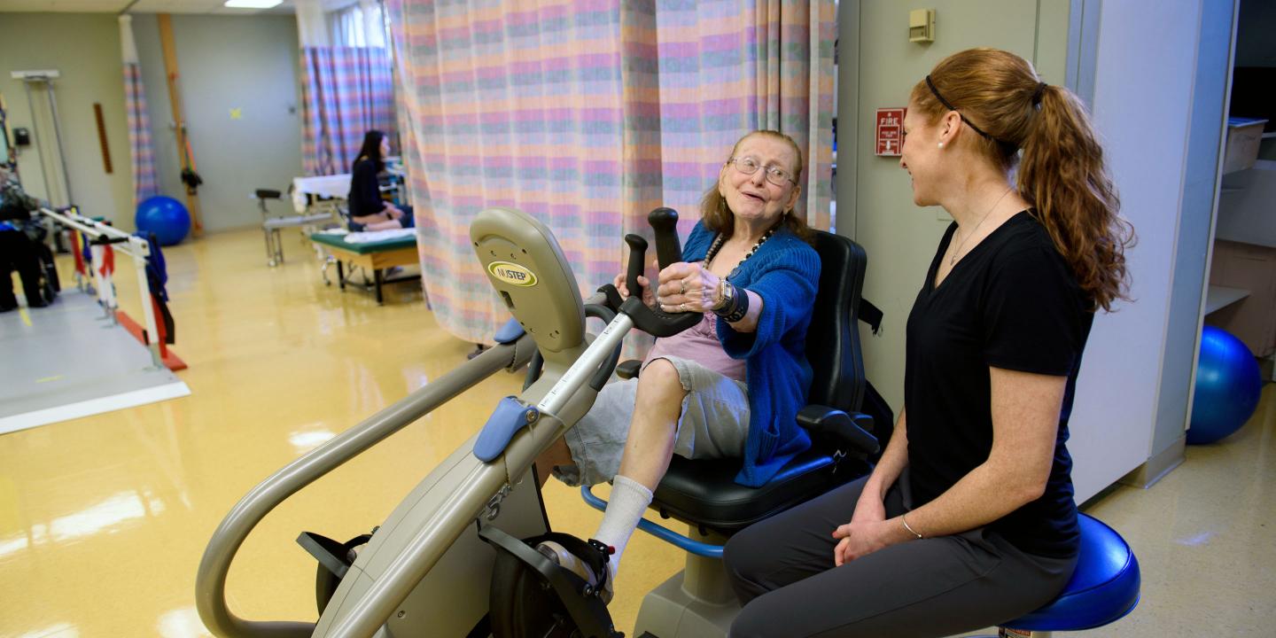 Physical therapist observes as an older woman uses a stationary bike in rehabilitation gym