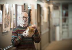 An older bearded man in a multi-color sweater standing in a hallway gallery at Orchard Cove holds a ceramic pot he has made.