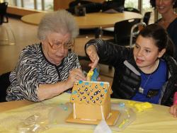 Older woman sits with a young girl as they put the finishing touches on a gingerbread house at Simon C. Fireman Community