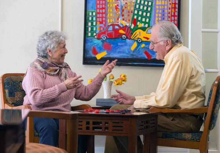 An older man and woman at Center Communities of Brookline play a game of checkers, near a brightly colored painting of city life.
