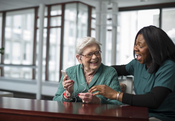 An older woman listens to an iPod. A staff caregiver has her arm around the woman. They're both laughing.