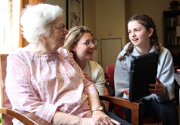 An eighth grader with braided pigtails holds up a tablet as she shares a conversation in Spanish with a memory care patient.