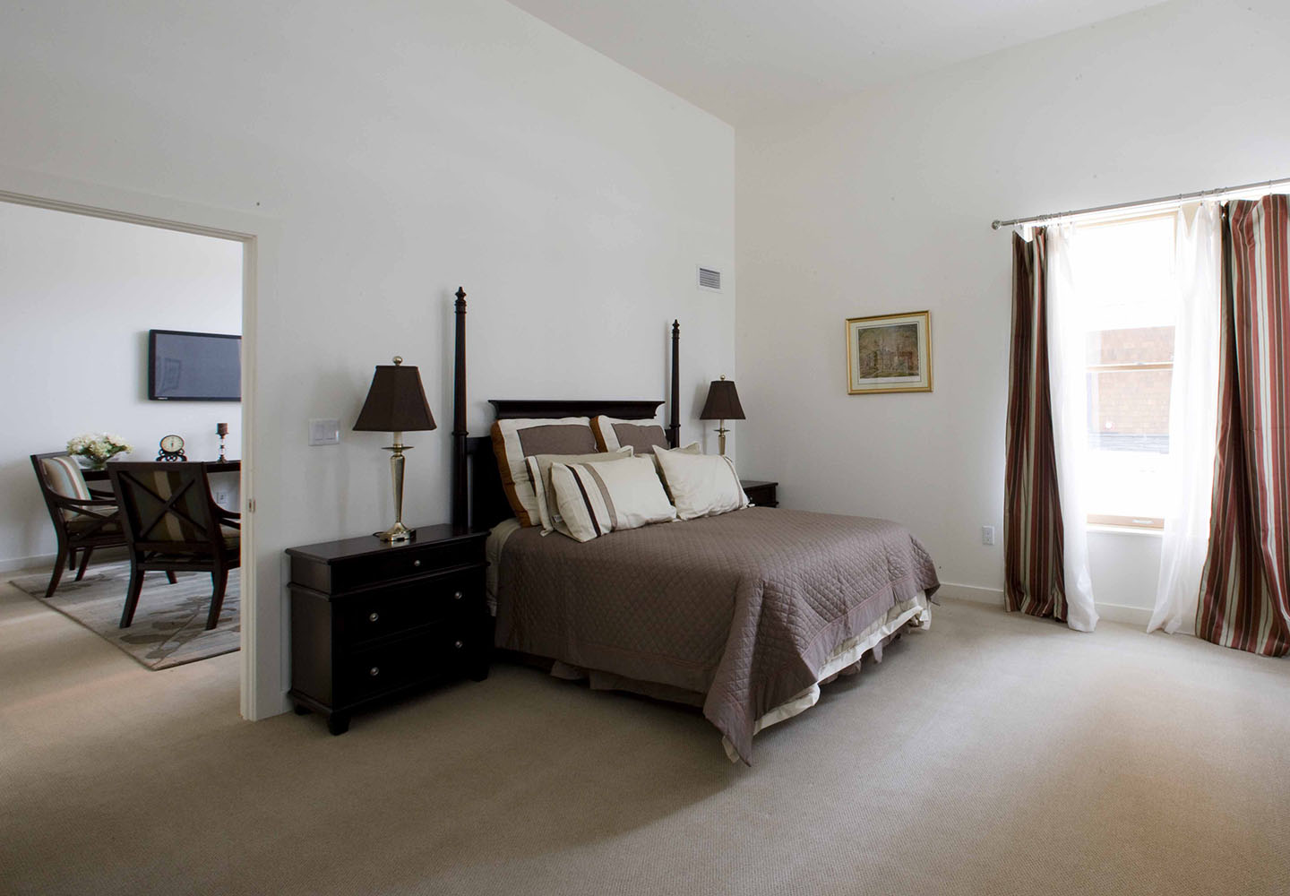 A bedroom with high ceilings and large windows opens to a spacious living room in an assisted living apartment at NewBridge on the Charles.