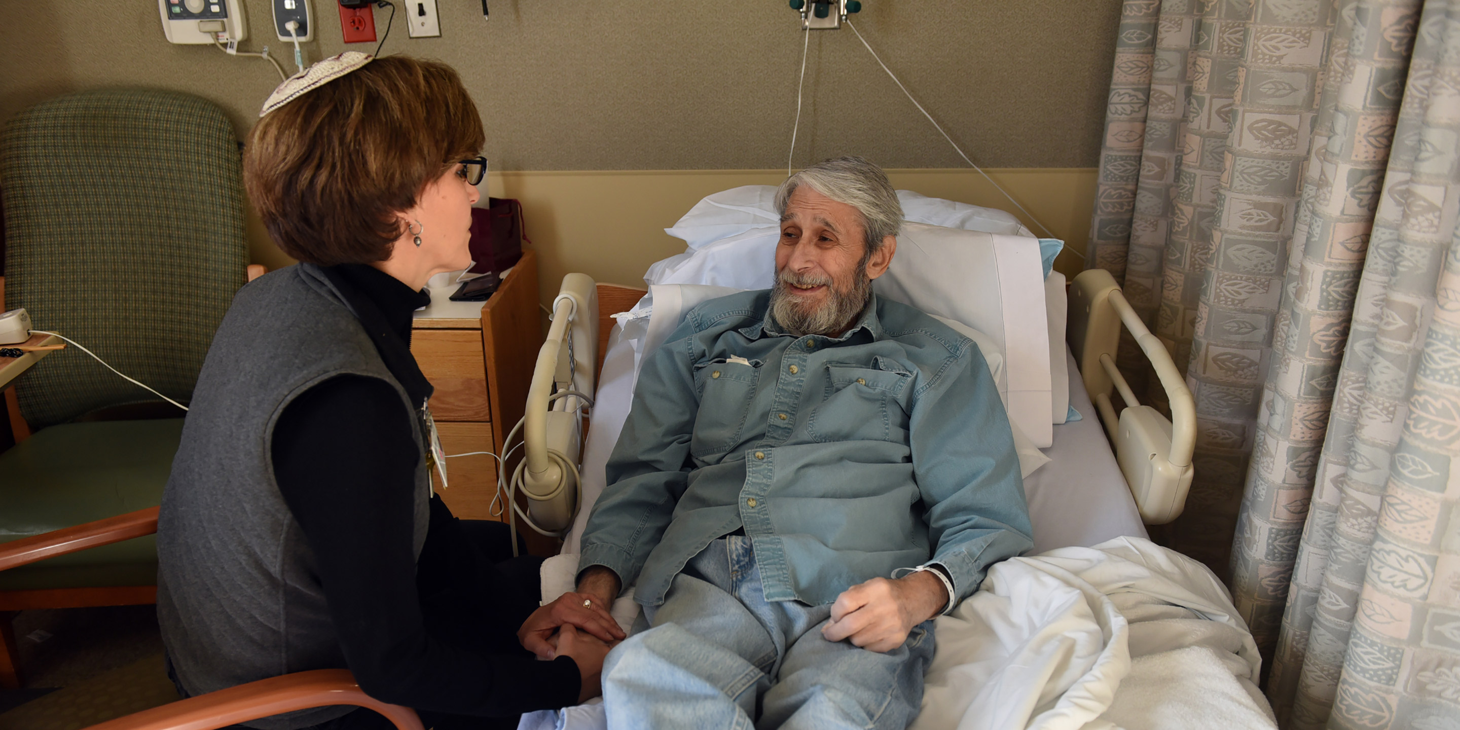 Rabbi Sara Paasche-Orlow sits and talks with a male patient while he lays in a hospital bed.