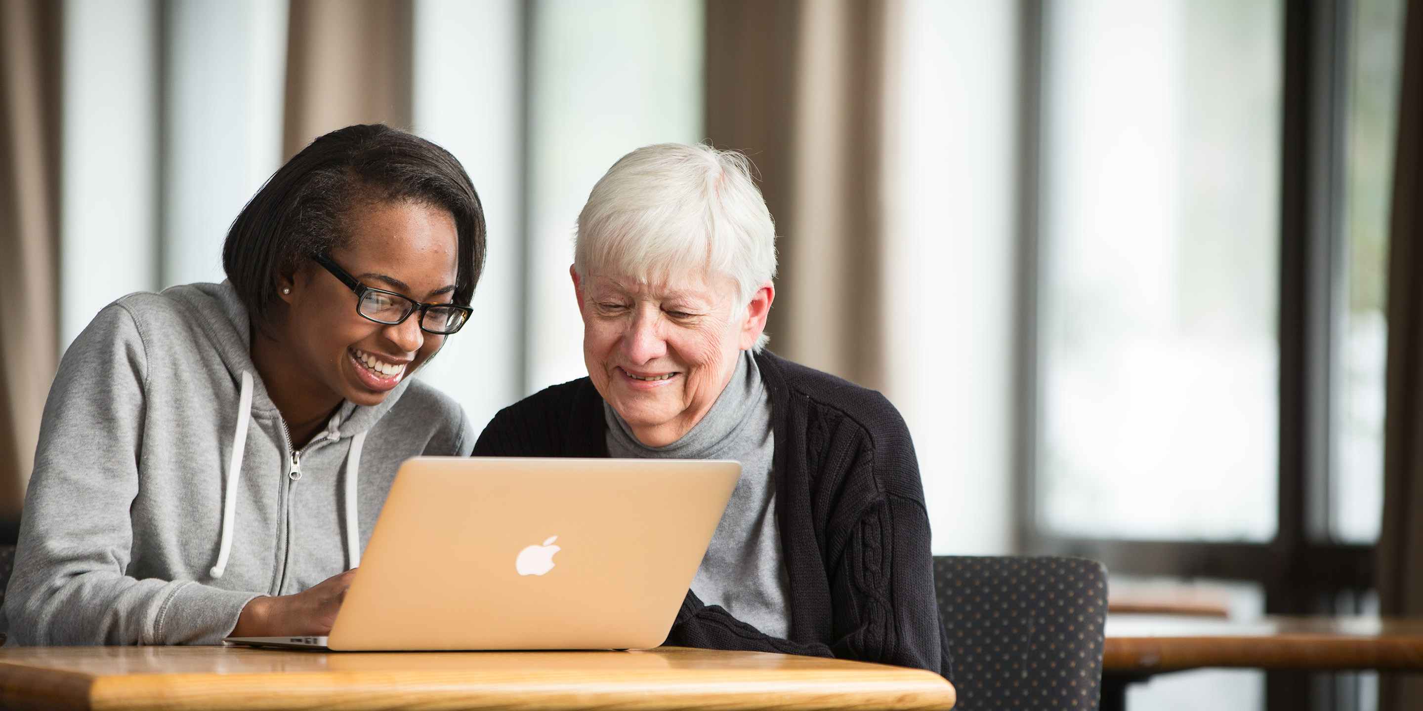 A female volunteer sits with an elder female and helps her use a laptop.