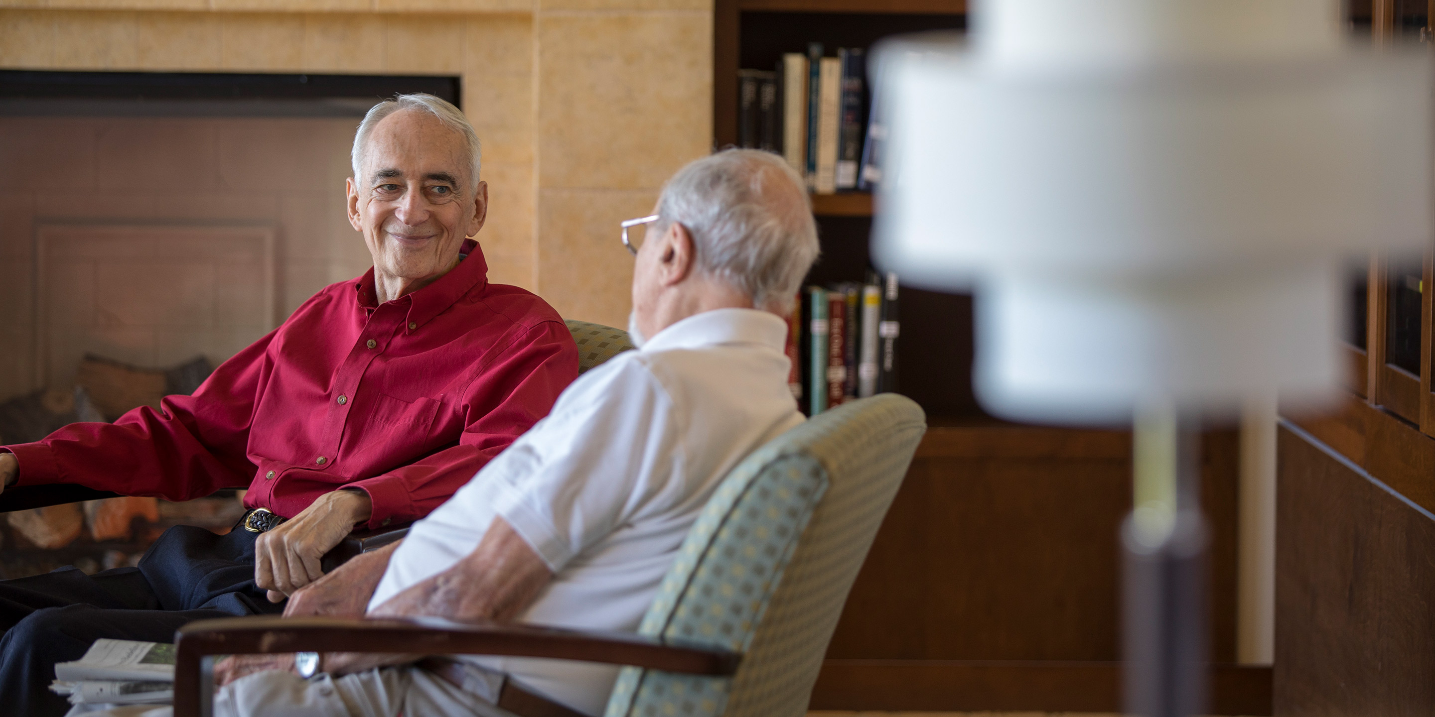 Two male residents sit in chairs in a library and talk to each other.