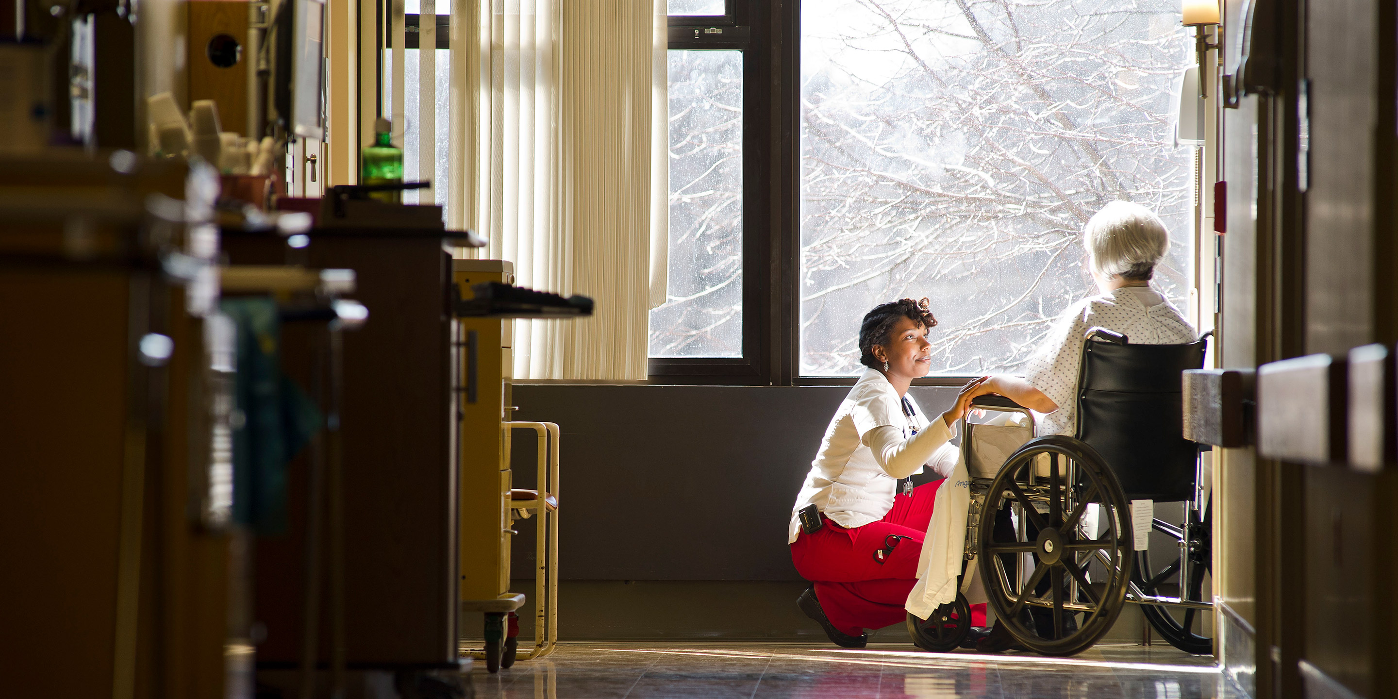 A female patient in a wheelchair is being comforted by a female caregiver who bends down to hold her hand 