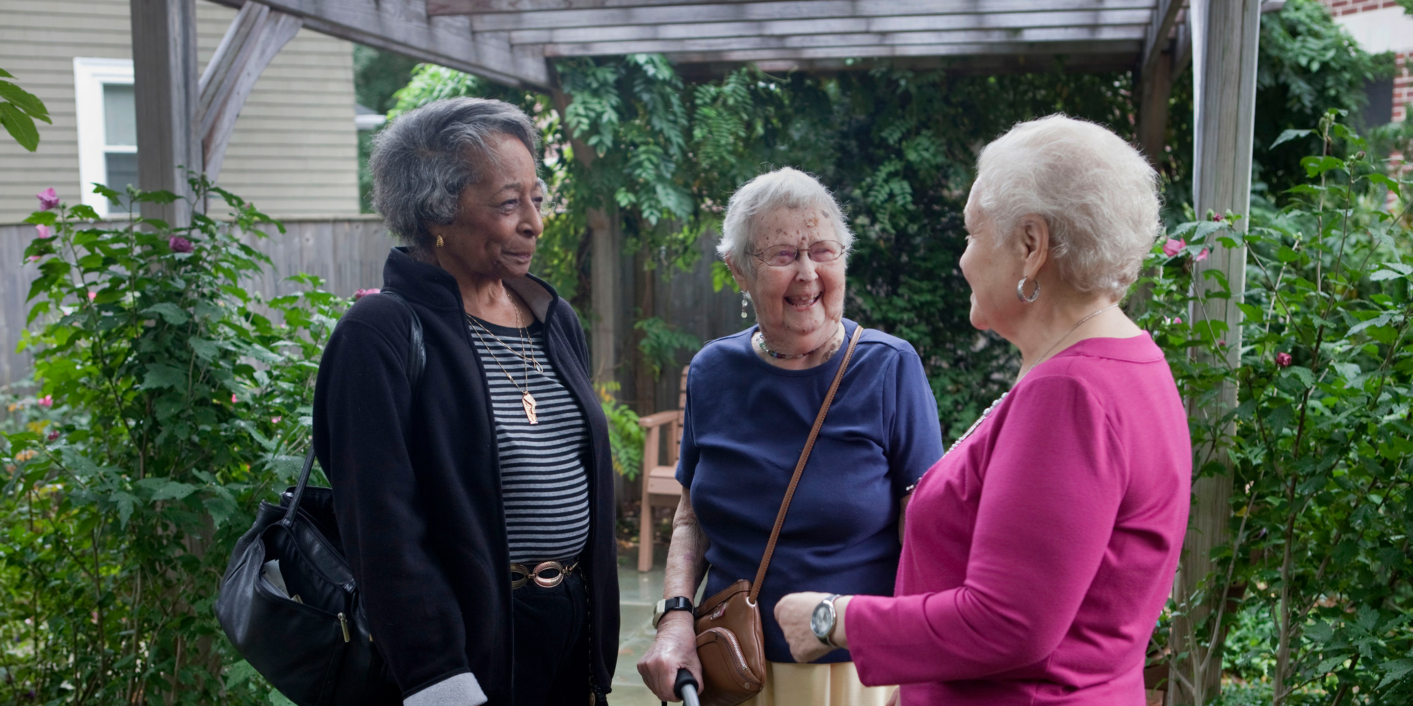 Three women stand together smiling outside surrounded by greenery 