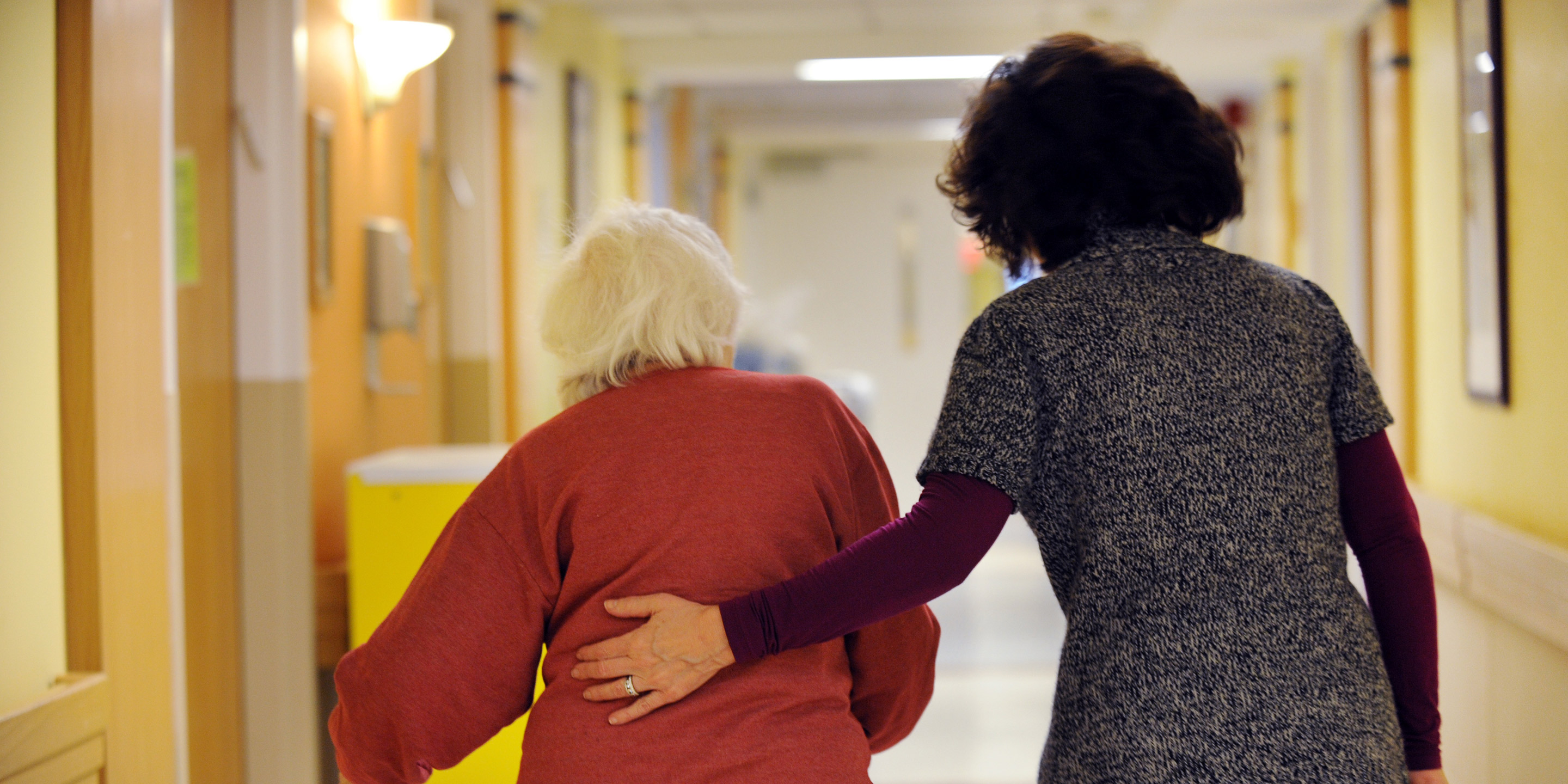 A woman with a walker walks down a hallway with another woman who helps guide her