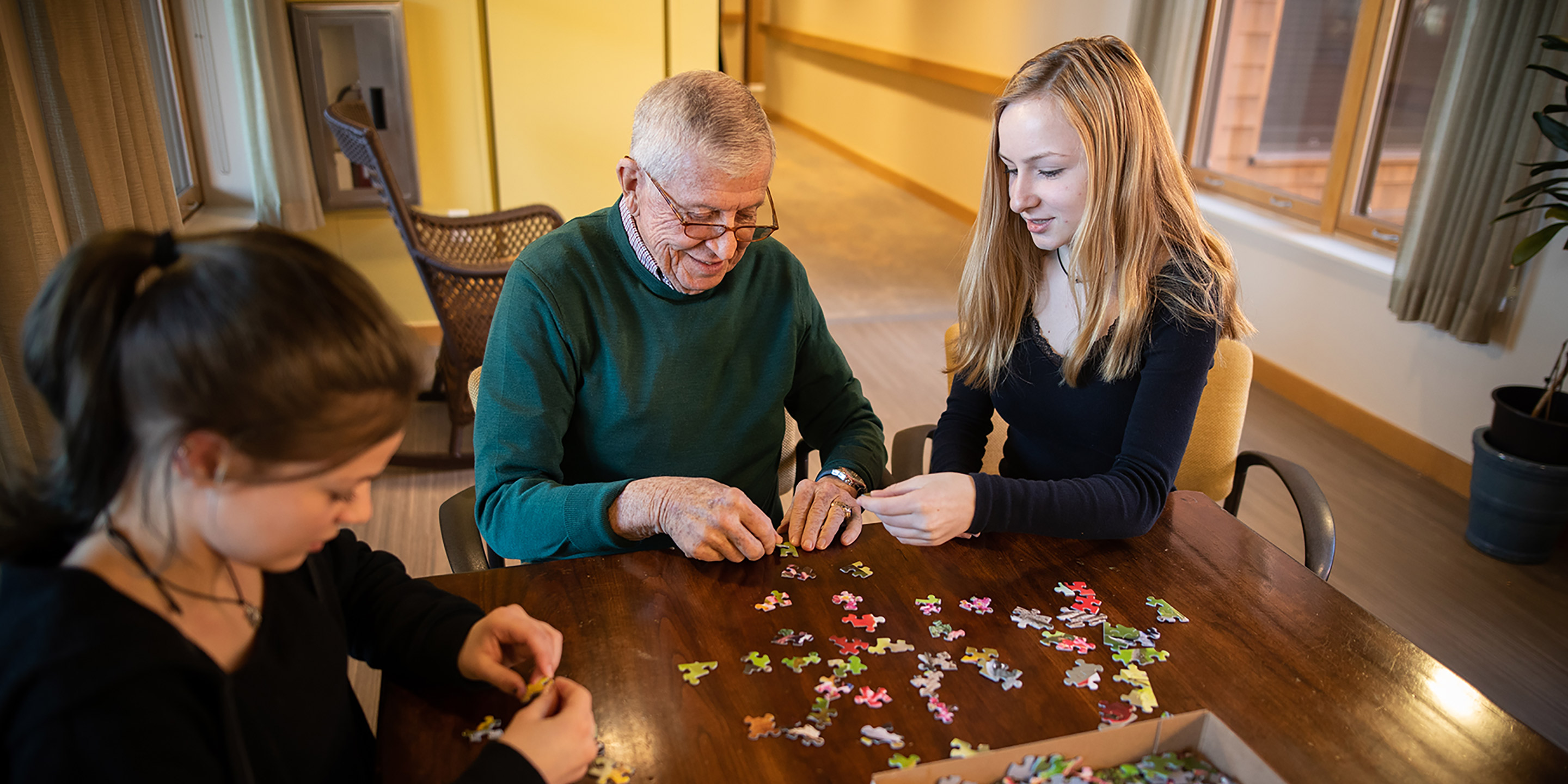 Two young girls sit with an elder man around a table and work on a puzzle together.