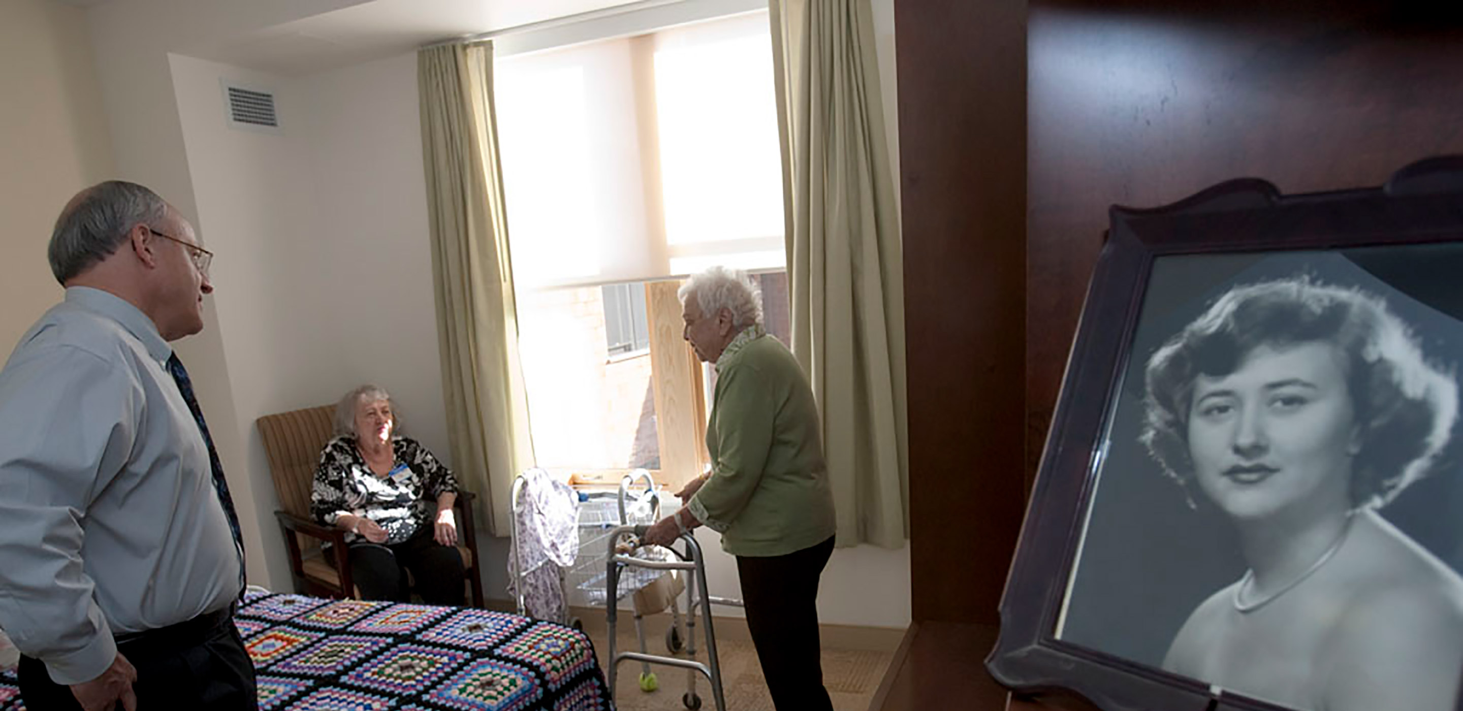 Hebrew SeniorLife's CEO Lou Woolf stands near a residents bed while talking to her and a female friend 