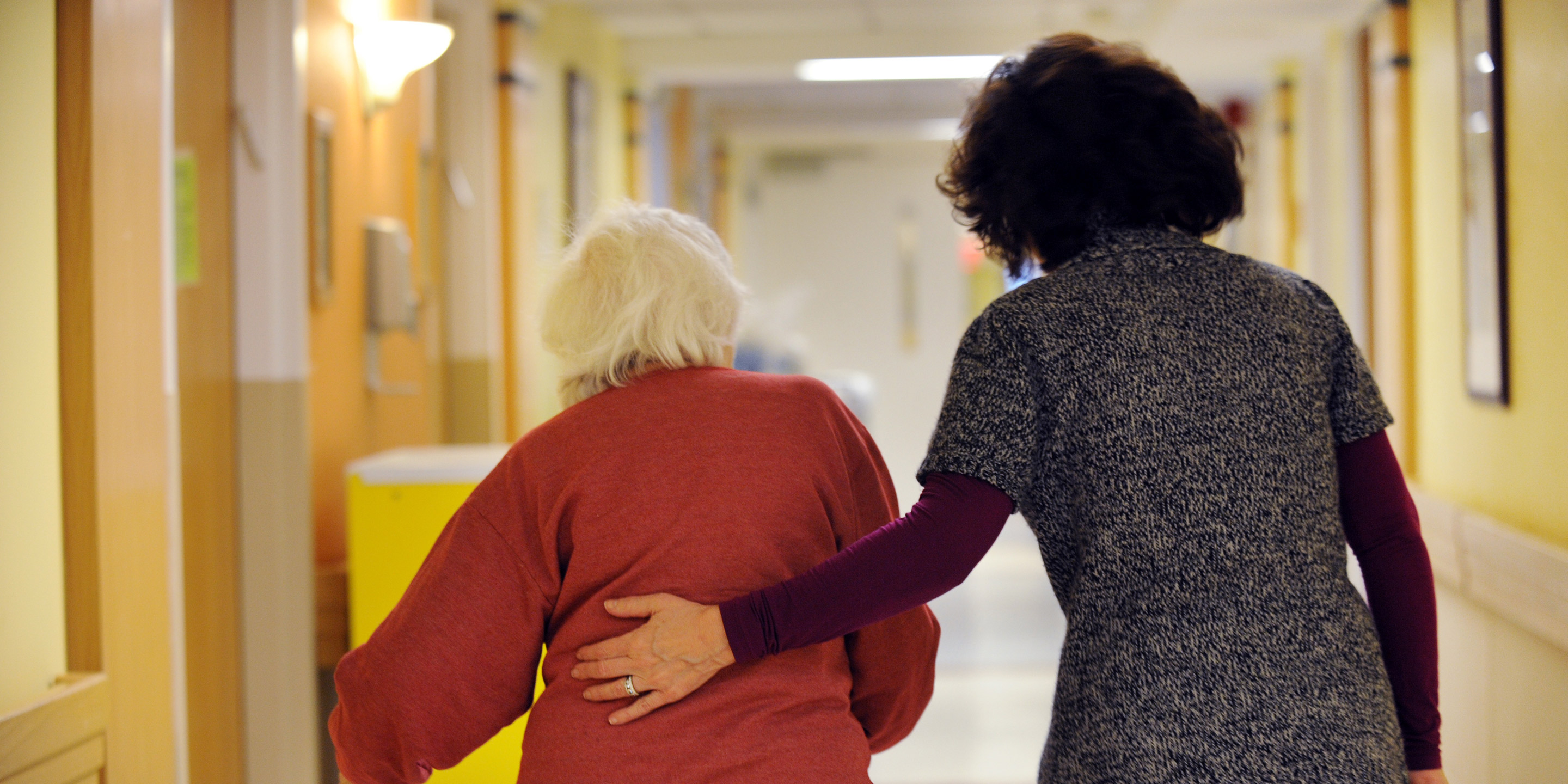 A woman with a walker walks down a hallway with another woman who helps guide her 