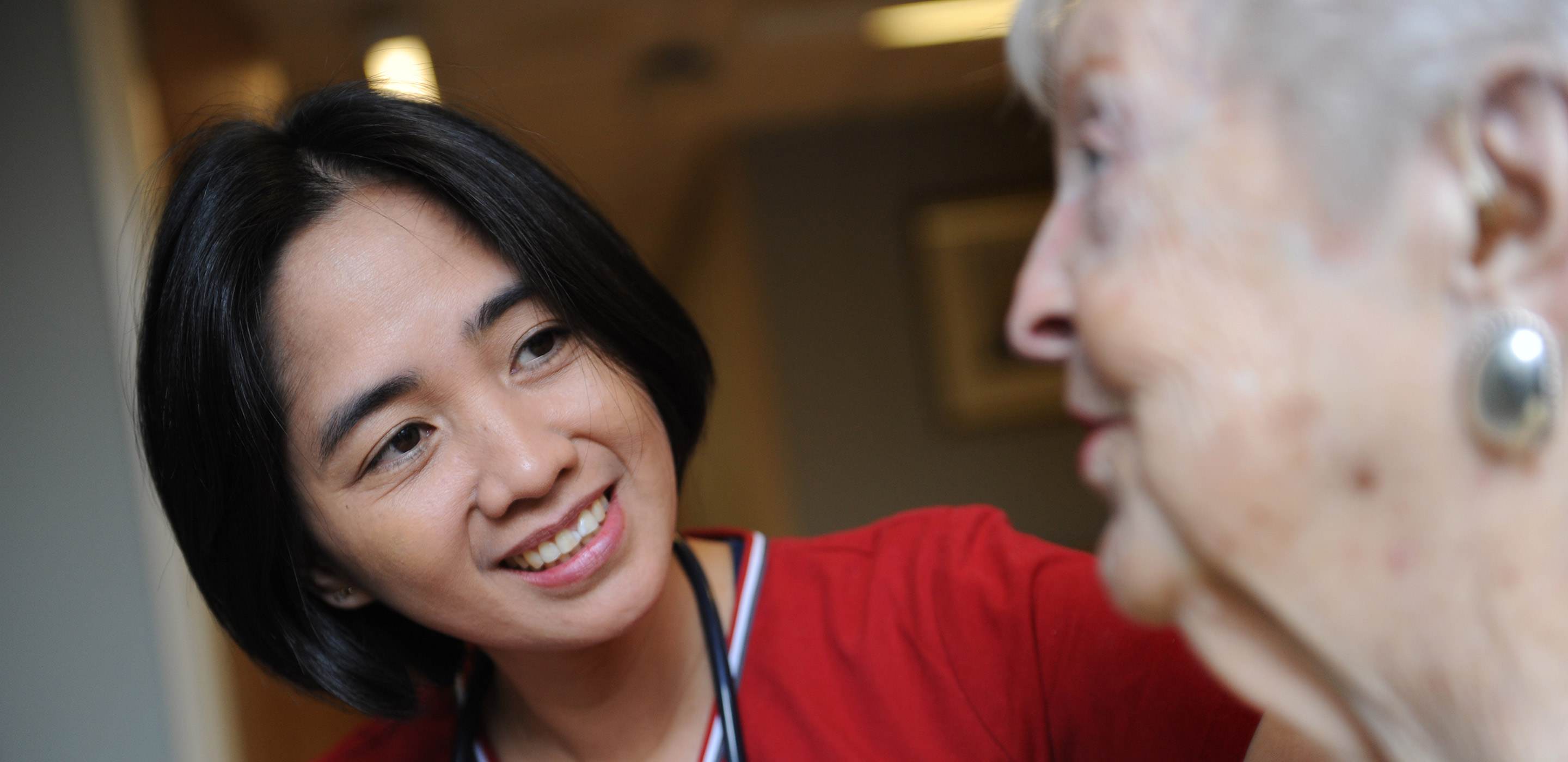 A female nurse in red scrubs sits and talks with a female resident 
