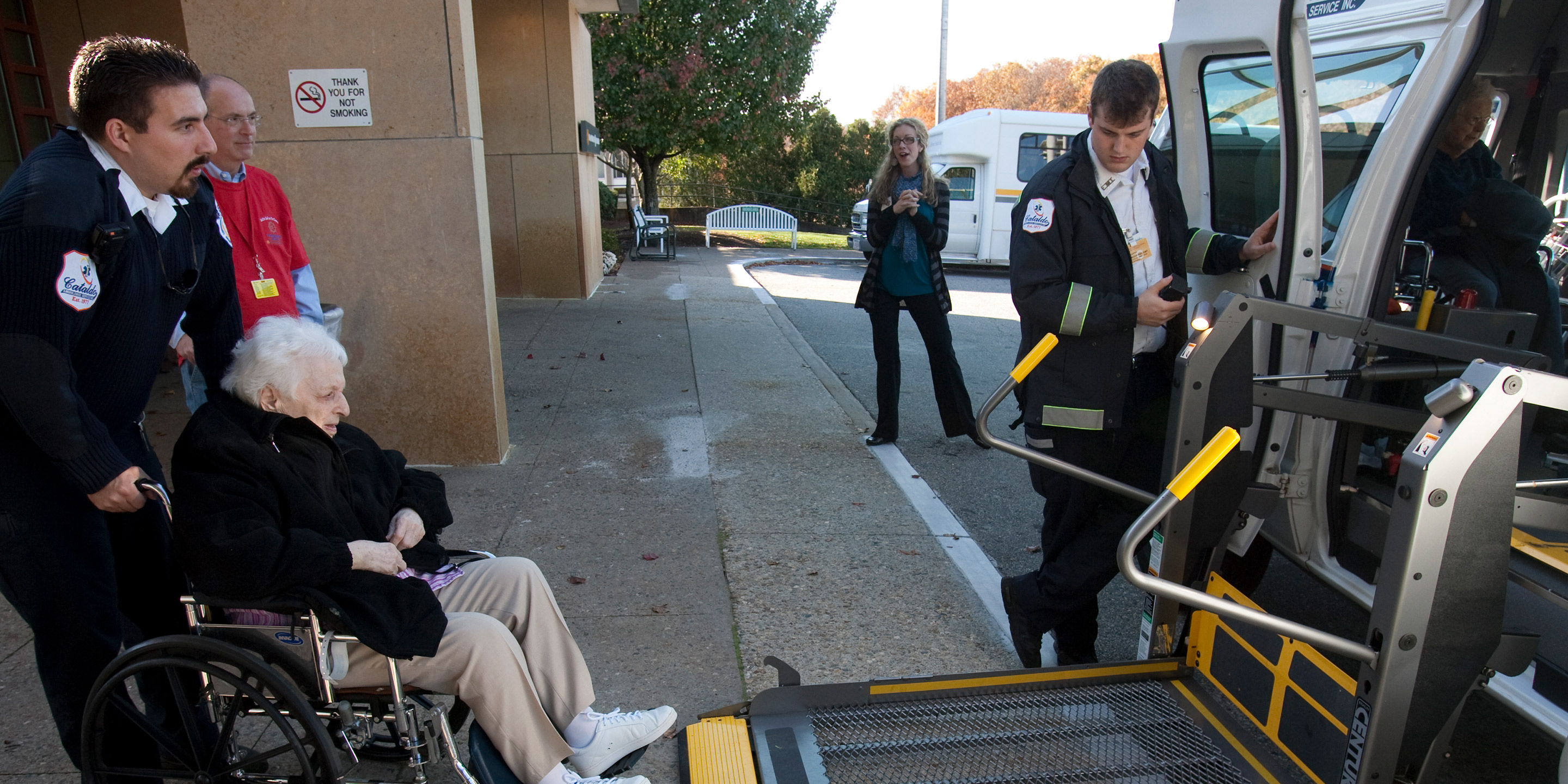 Two employees who work in the Security department at Hebrew SeniorLife help a resident in a wheelchair get loaded onto a lift to get into a transport vehicle 