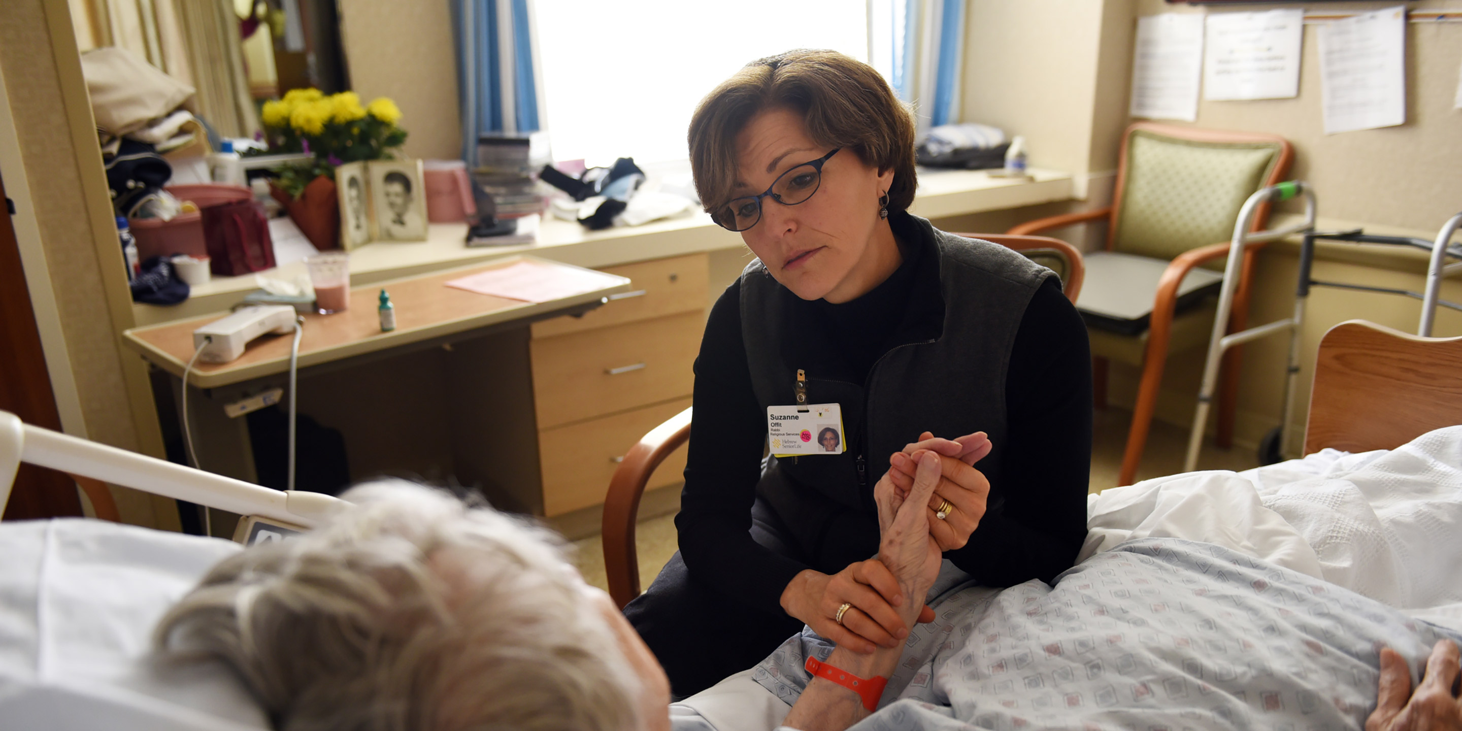 Rabbi Sara Paasche-Orlow  sits in a chair and holds a patients hand while talking to her 