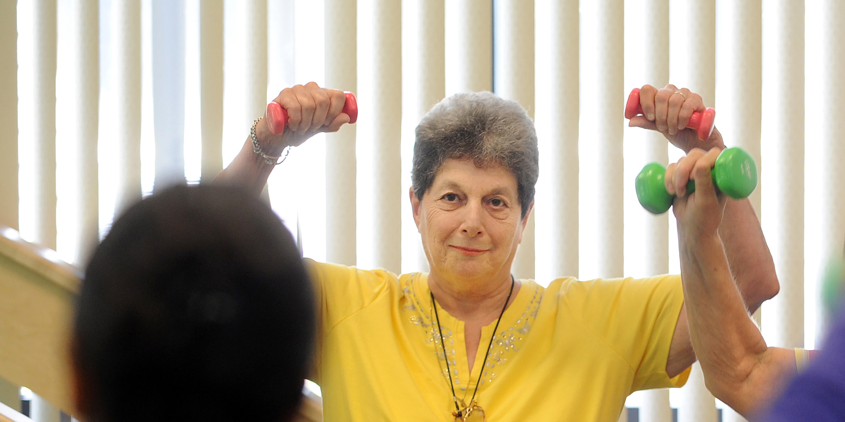 A female senior smiles and lifts dumbbells over her head in a fitness class