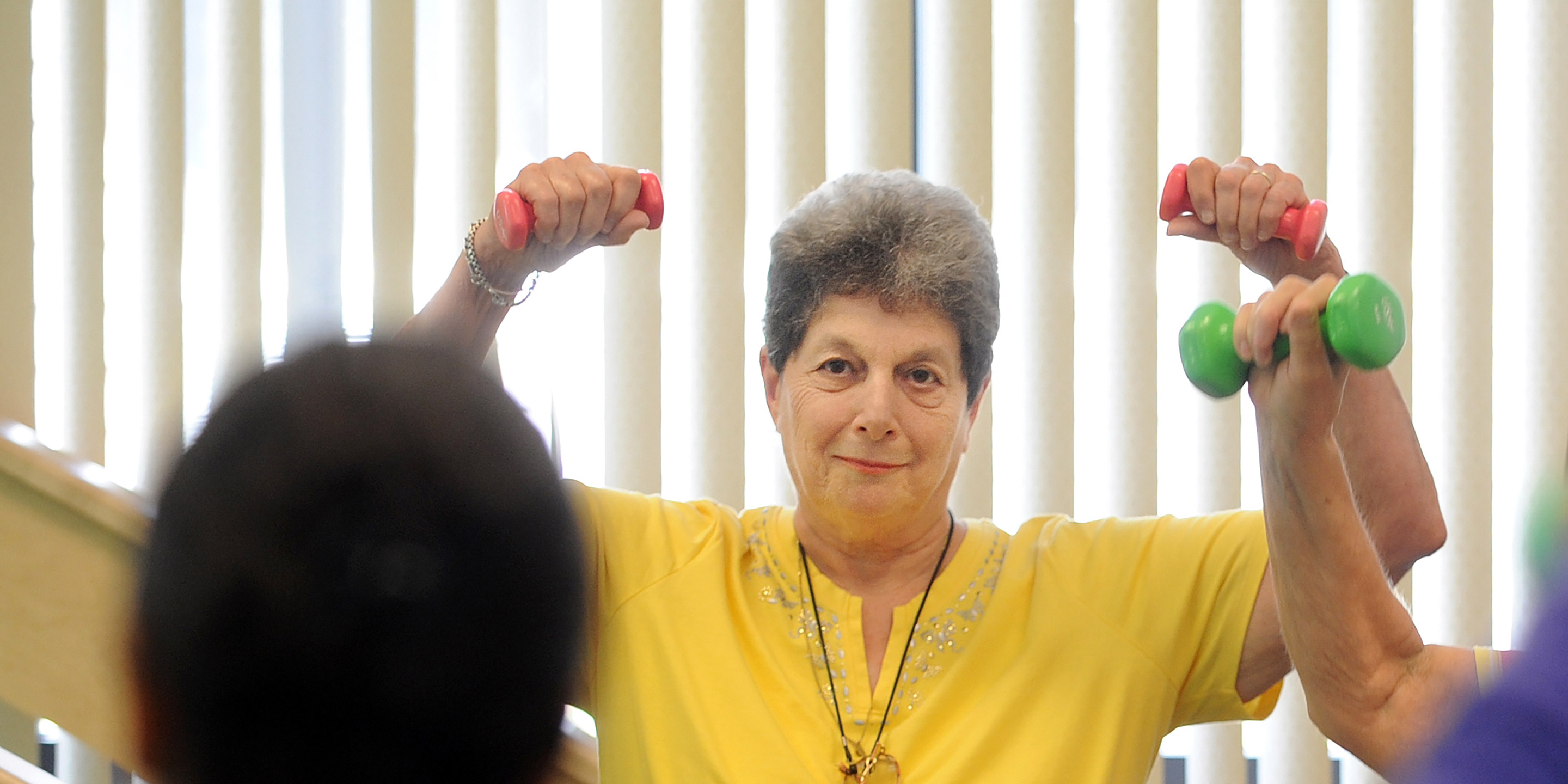 A female senior smiles and lifts dumbbells over her head in a fitness class 