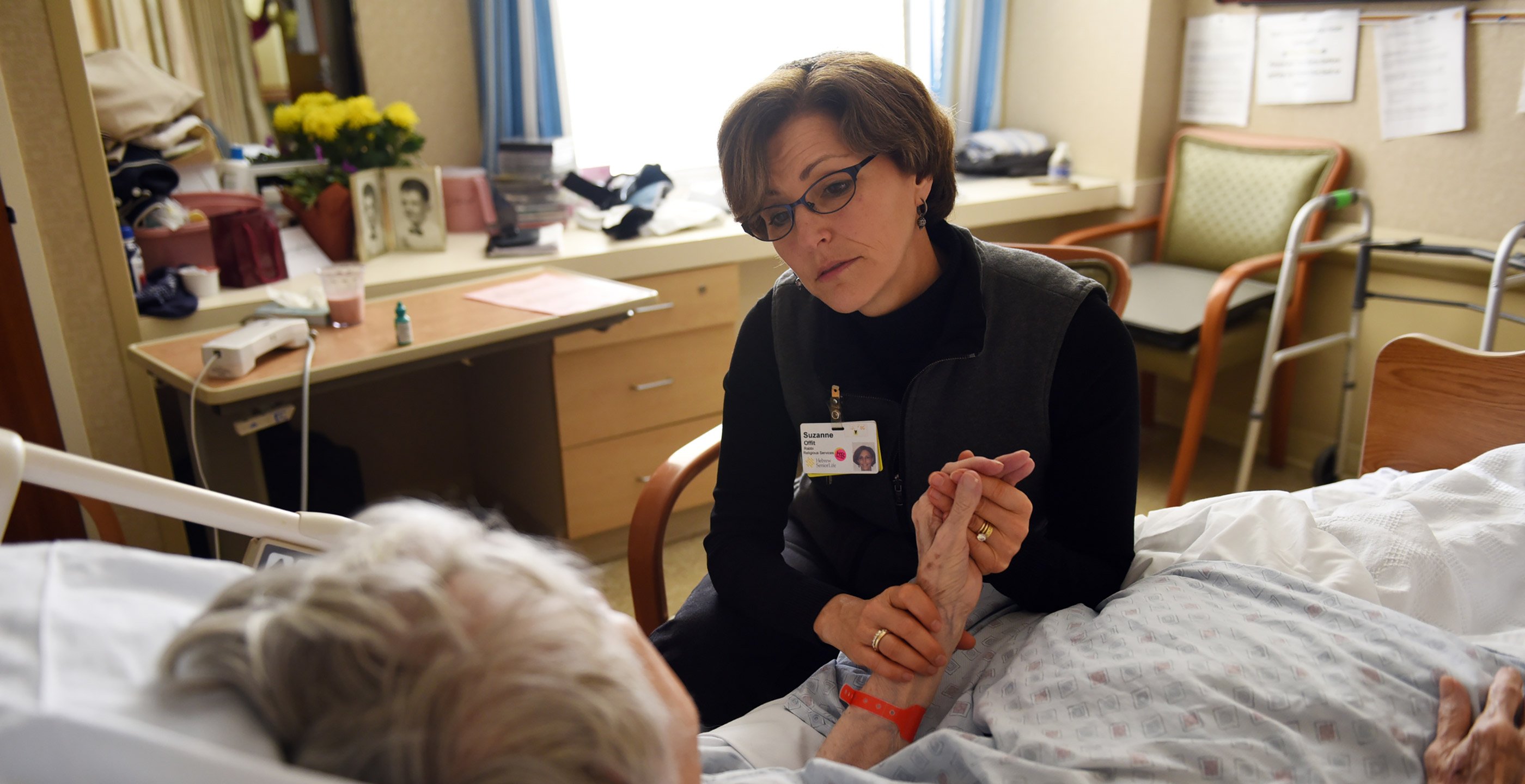 Rabbi Sara Paasche-Orlow  sits in a chair and holds a patients hand while talking to her 