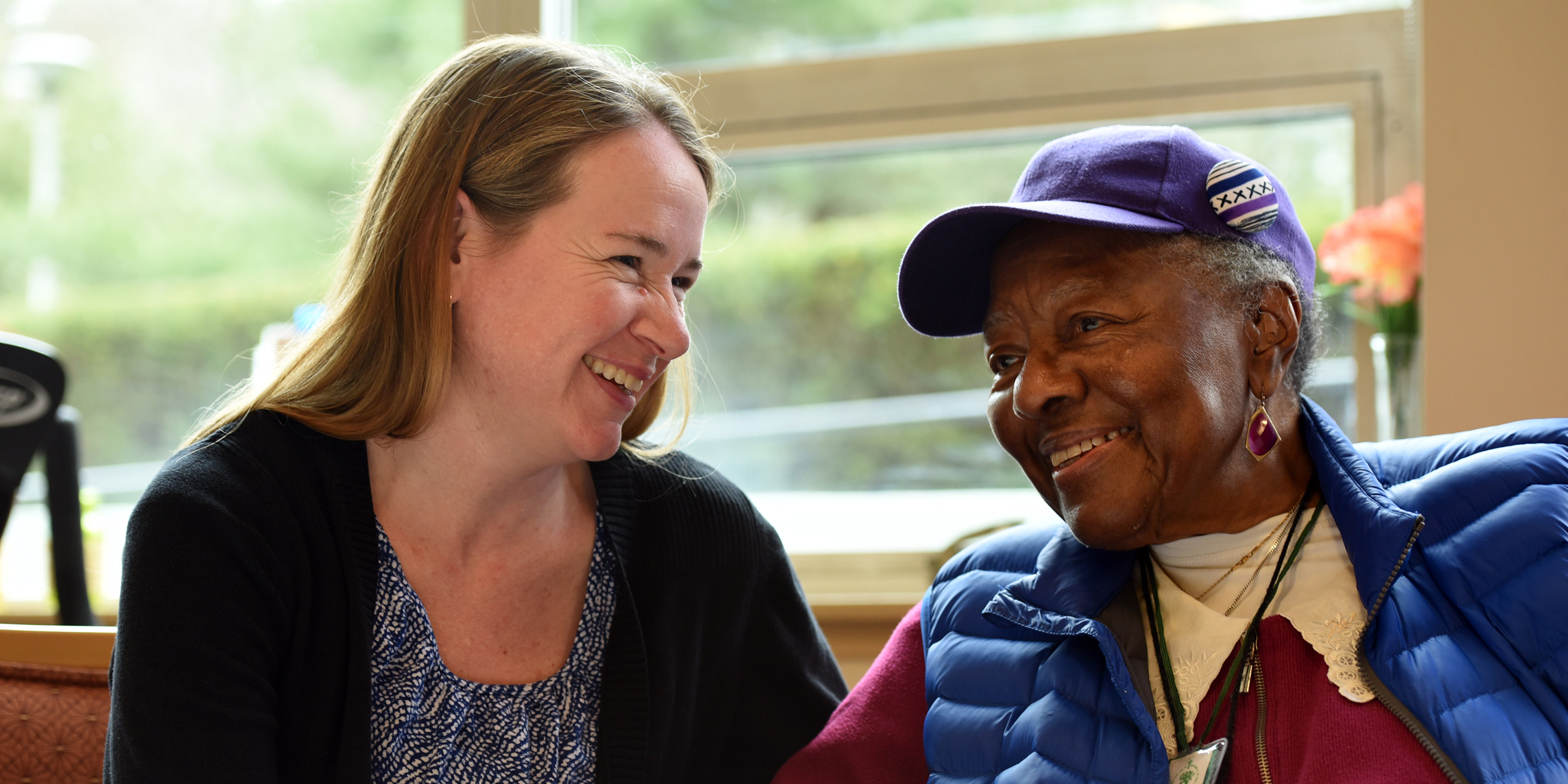 A Home Health worker smiles while sitting next to a smiling female senior