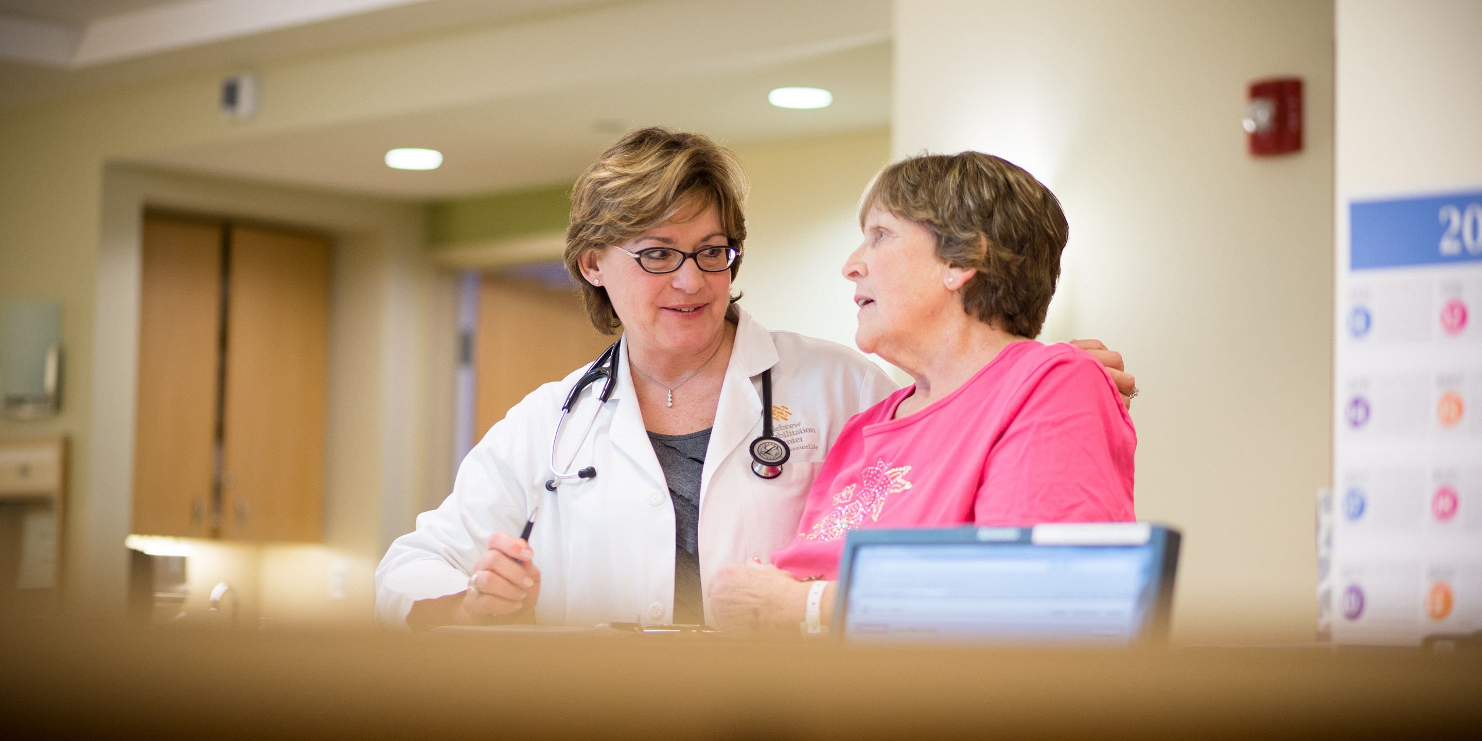 A Hebrew SeniorLife doctor puts her arm around a woman as she stands and talks with her 