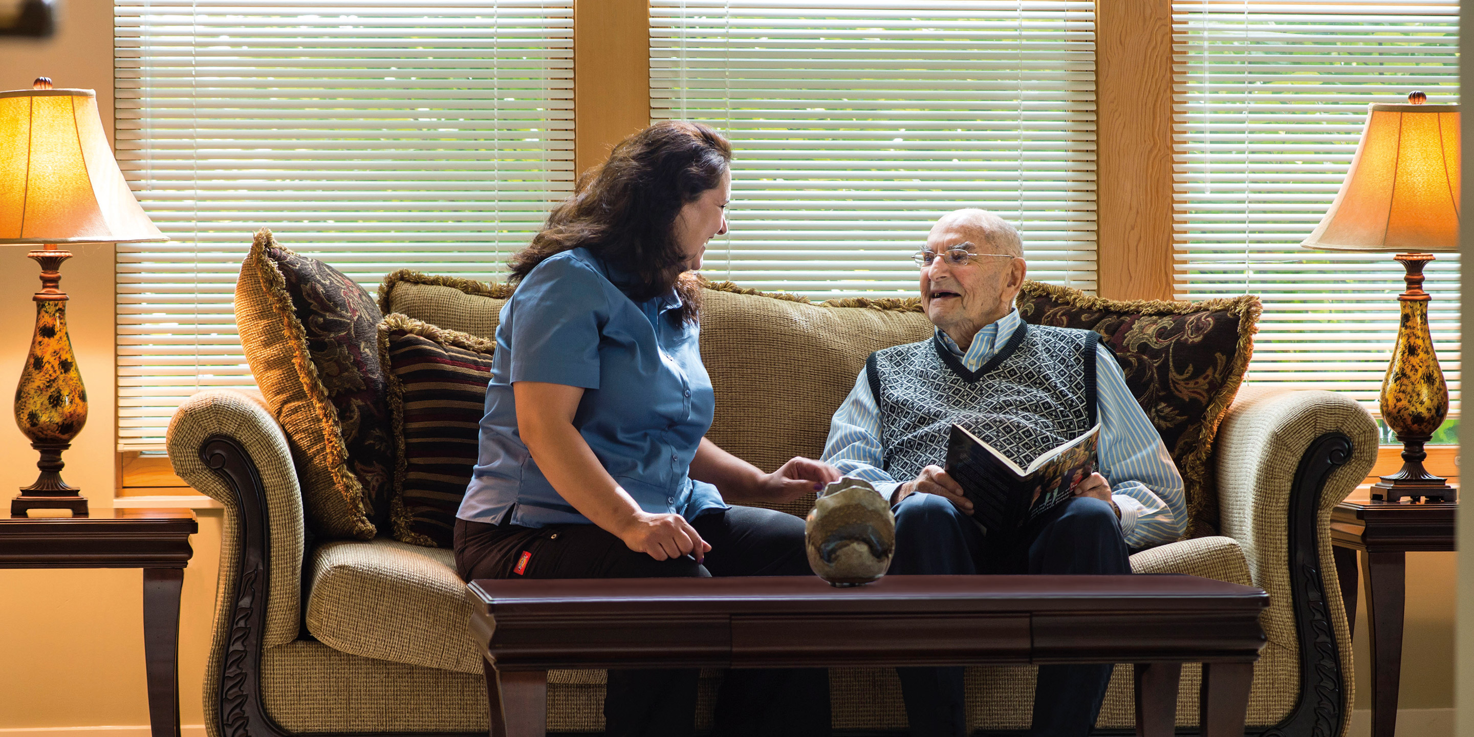 An older male sits on a couch with a woman caregiver and reads.