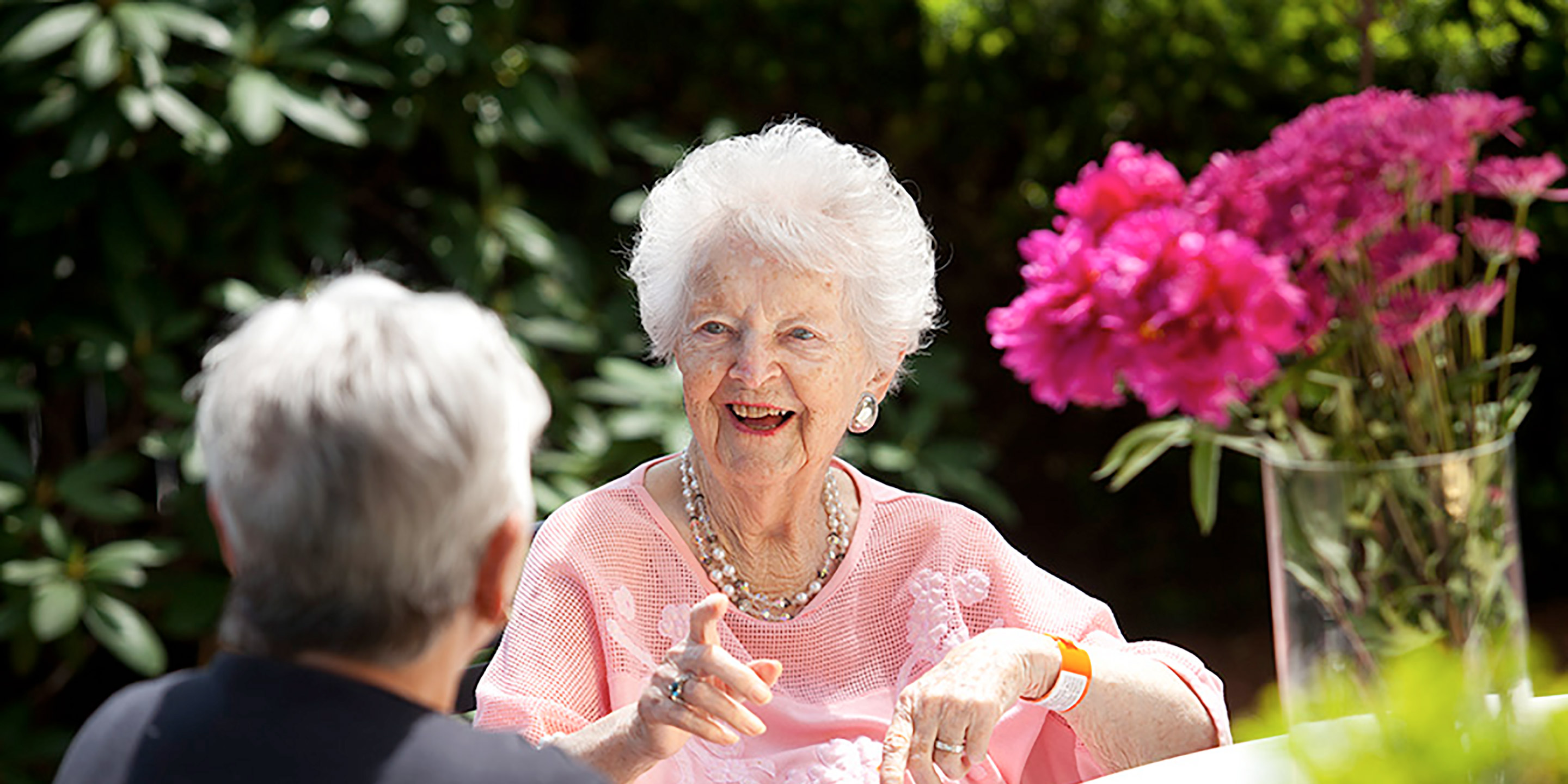 An elder woman wearing a pink shirt sits around a table outside and talks with another woman.