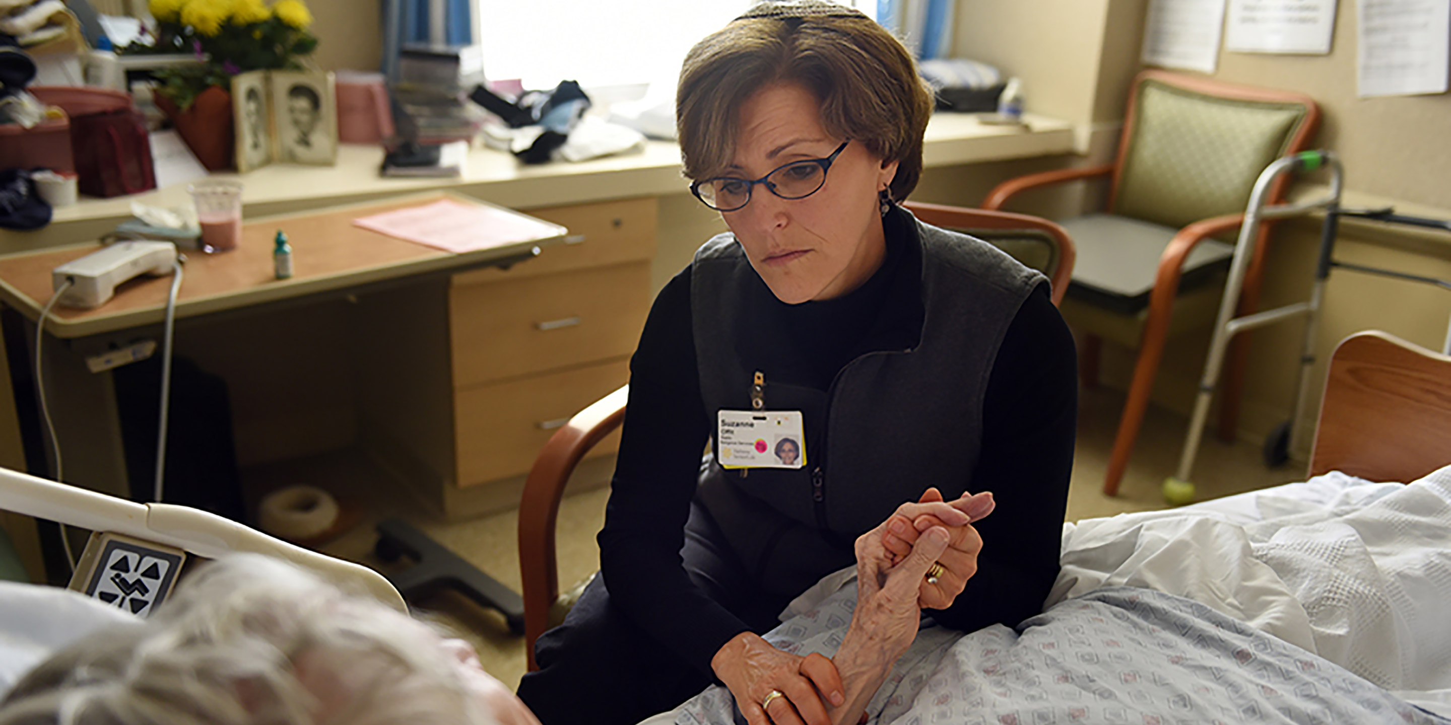 Rabbi Sara Paasche-Orlow  sits in a chair and holds a patients hand while talking to her 