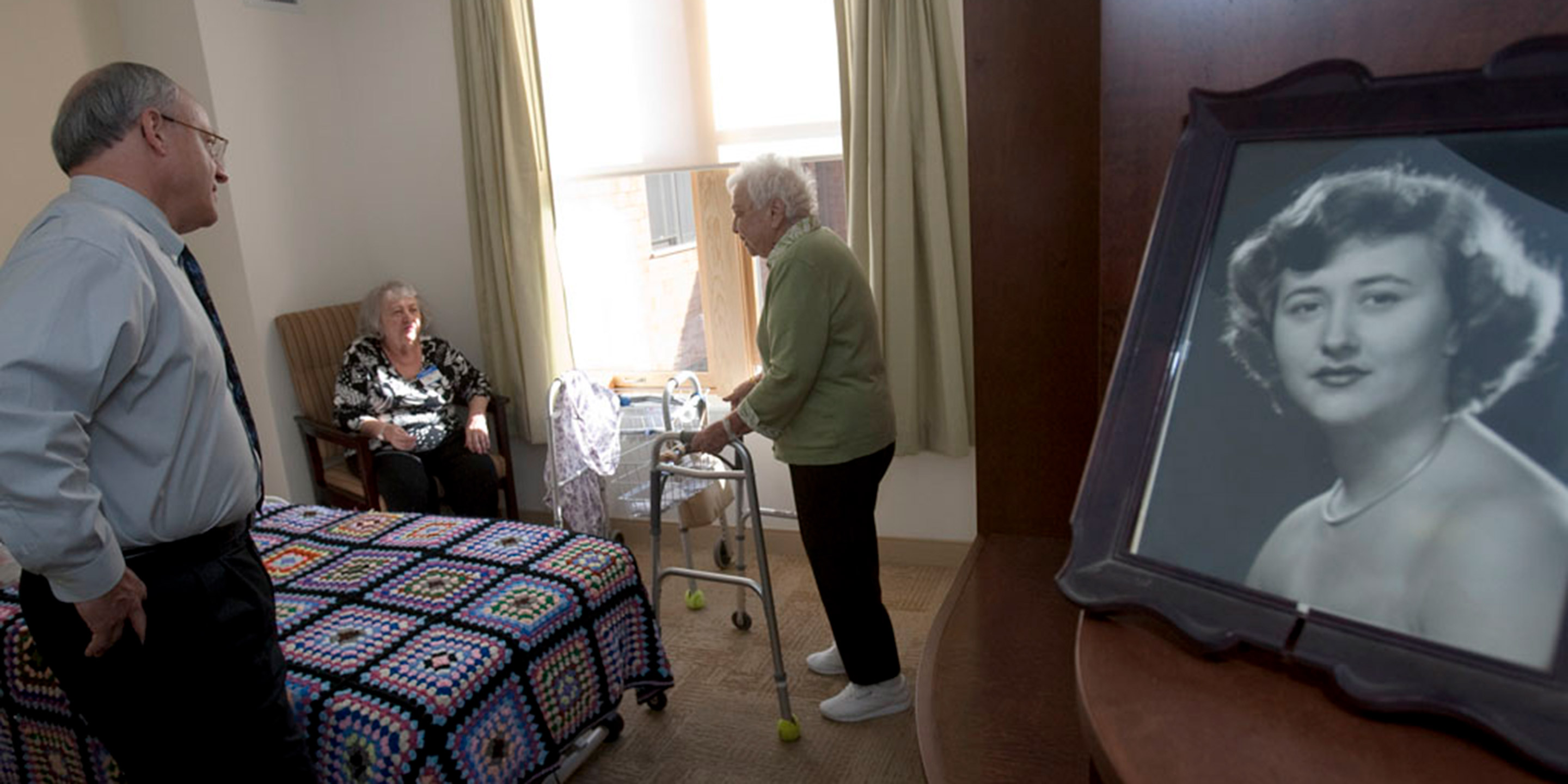 Hebrew SeniorLife's CEO Lou Woolf stands near a residents bed while talking to her and a female friend 