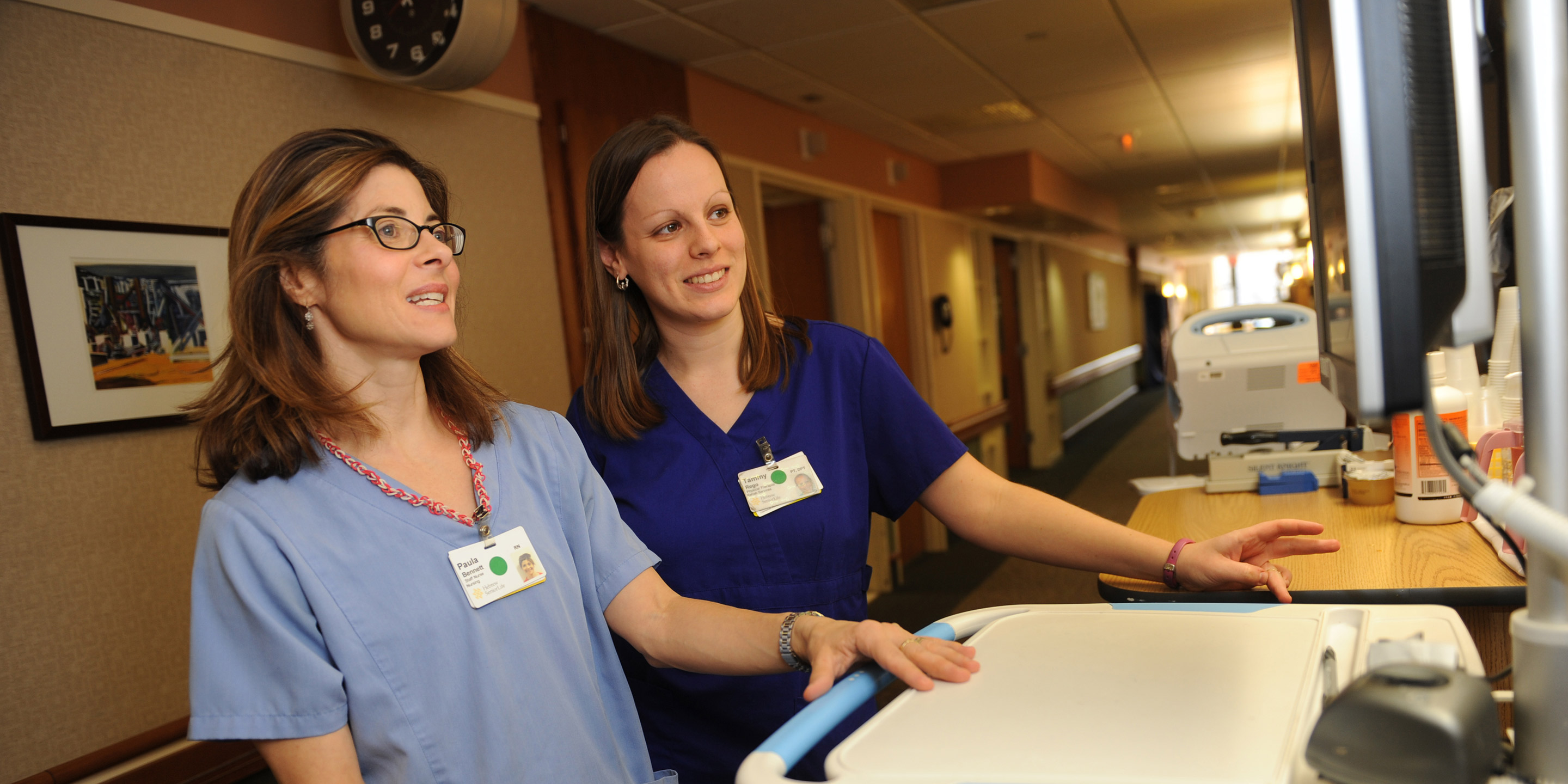 Two female nurses wearing blue scrubs look together at a chart on the computer