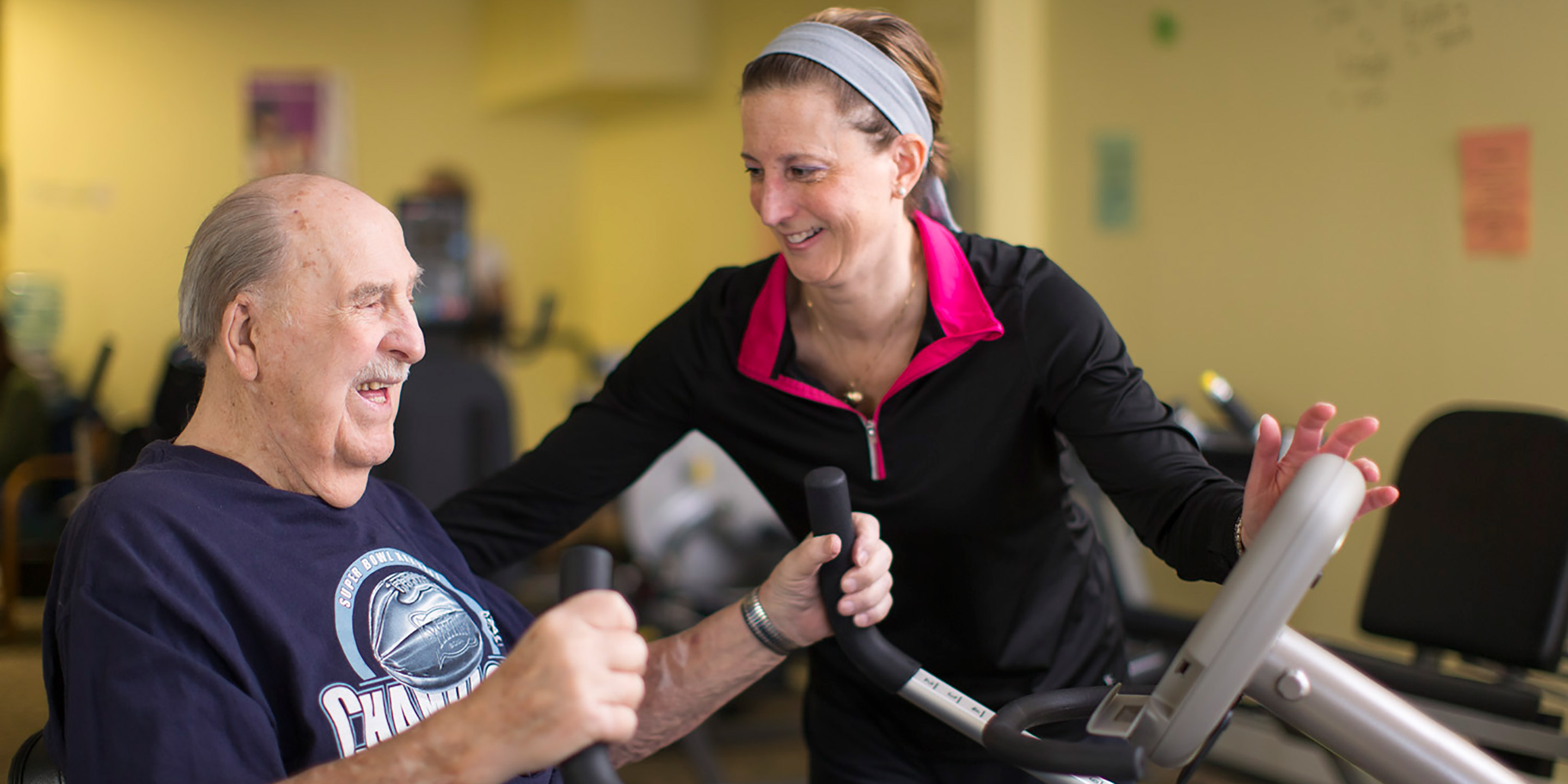 A female trainer helps an elder male while he works out on a stationary workout bike