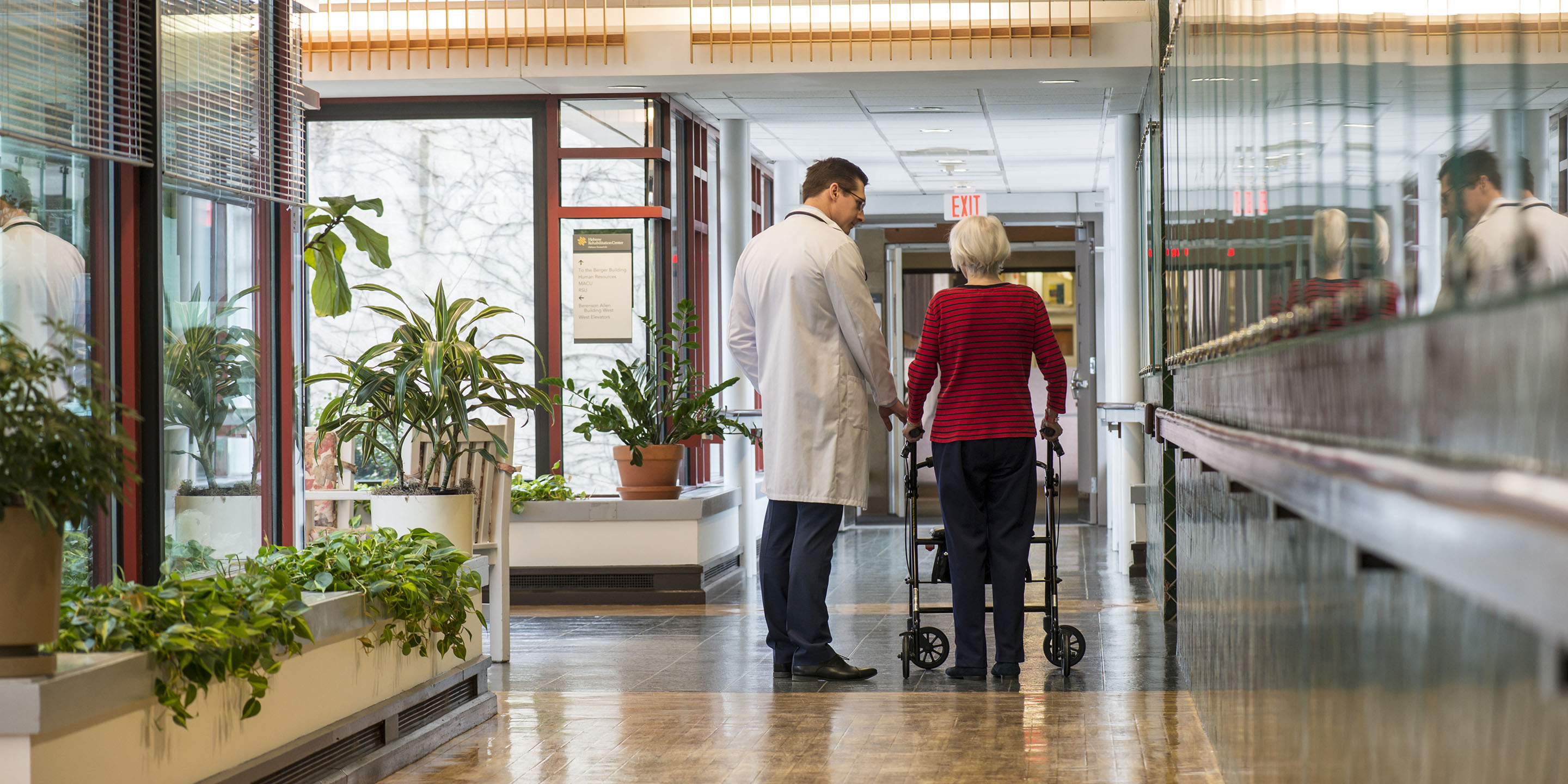 A male doctor talks with older woman using walker in Hebrew Rehabilitation Center hallway