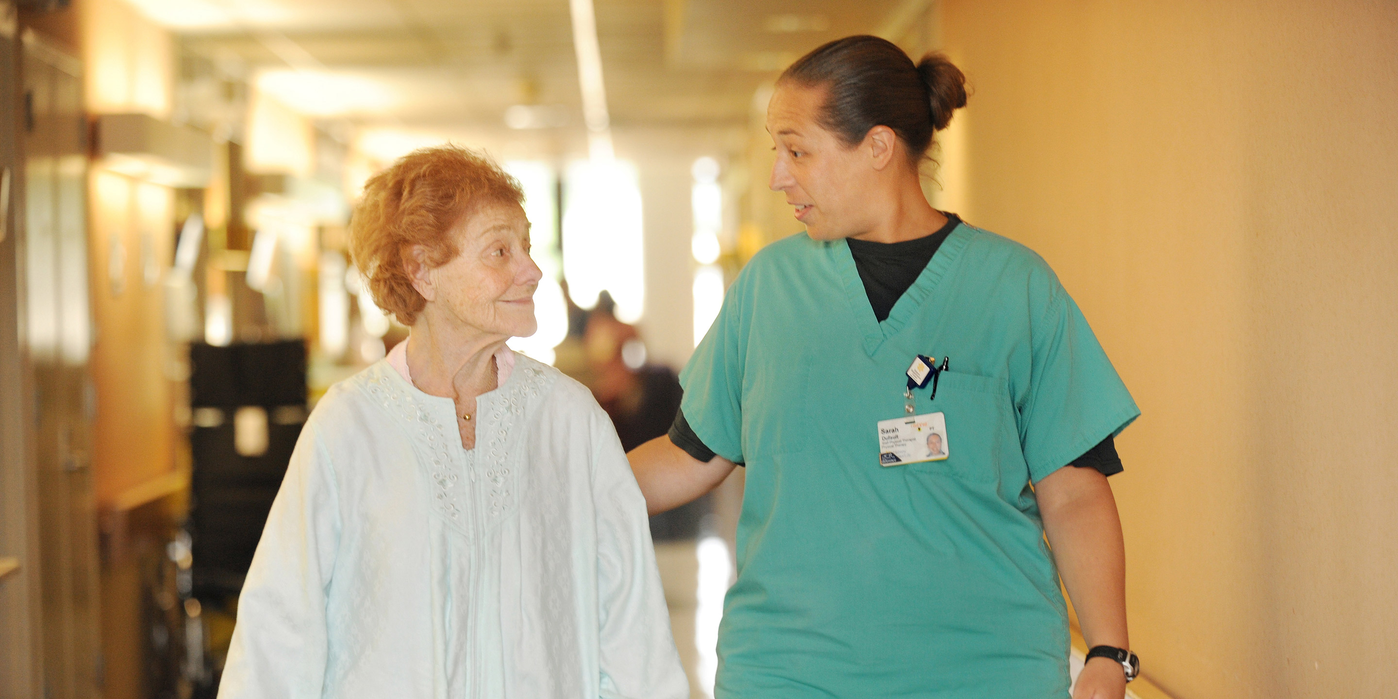 A patient smiles while walking down a hallway with a staff member at HRC Roslindale.