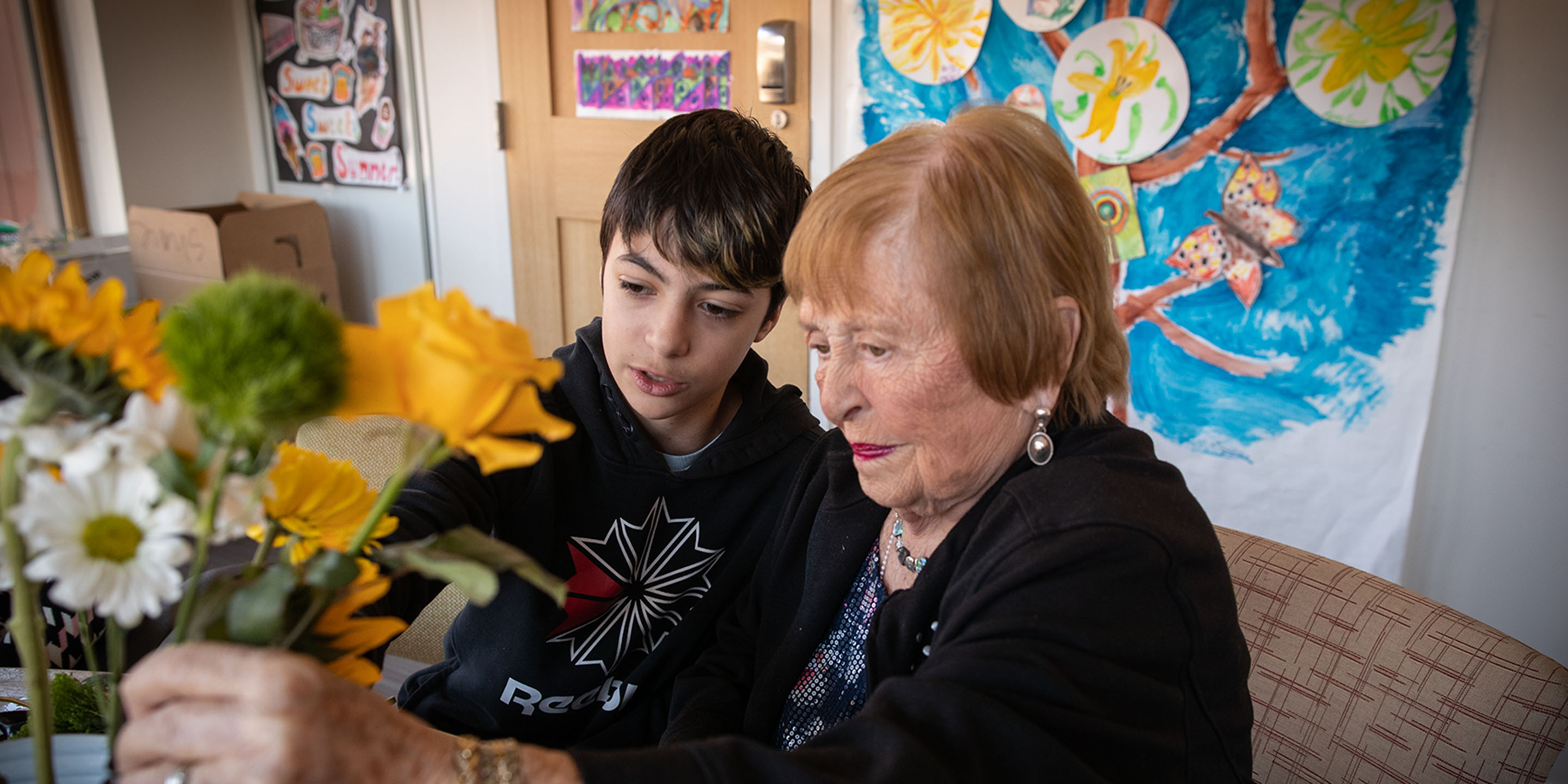 A resident in memory assisted living at NewBridge on the Charles in Dedham, MA arranges flowers with a middle-school aged student.