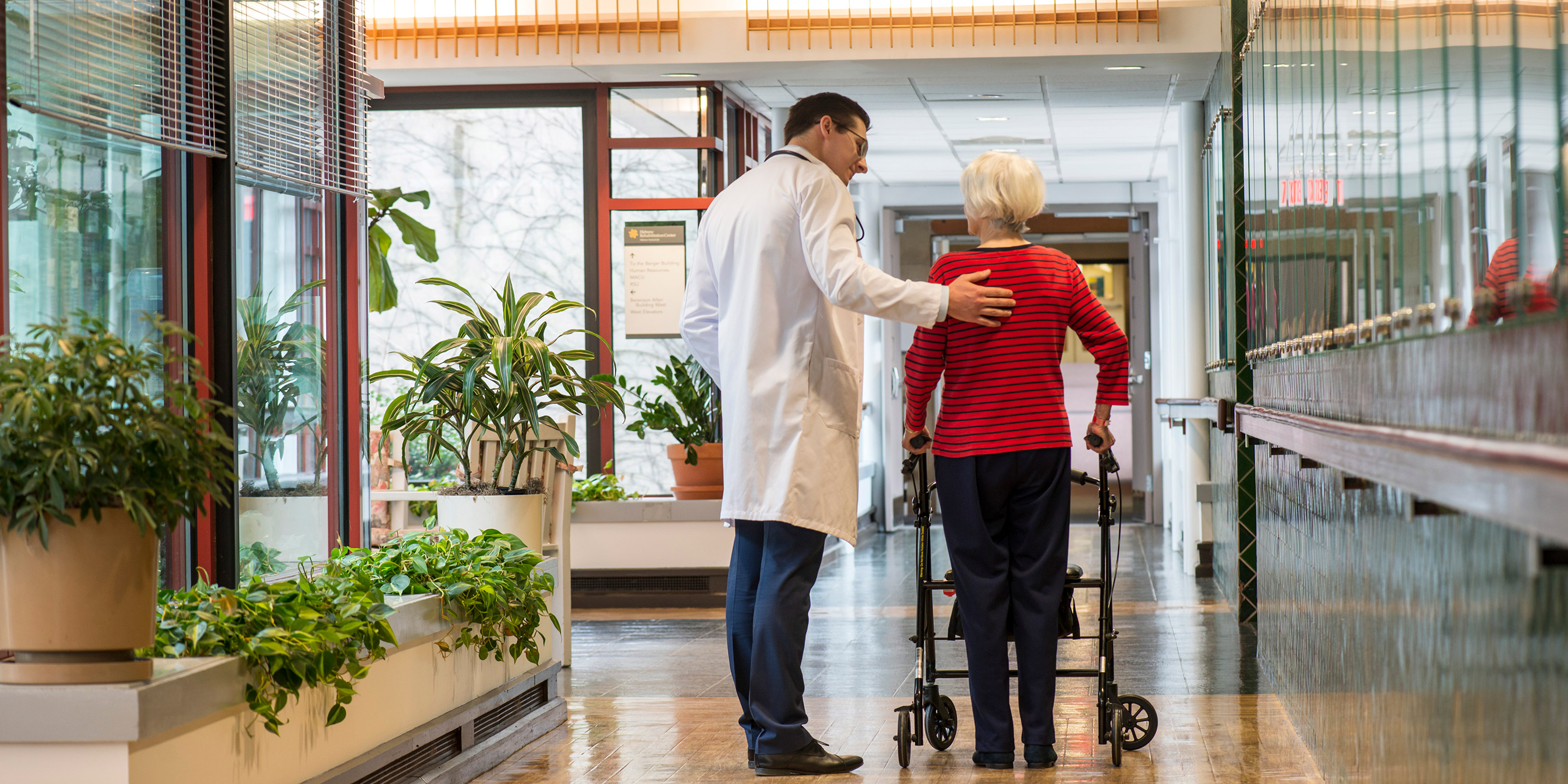 A male doctor talks with older woman using walker in Hebrew Rehabilitation Center hallway