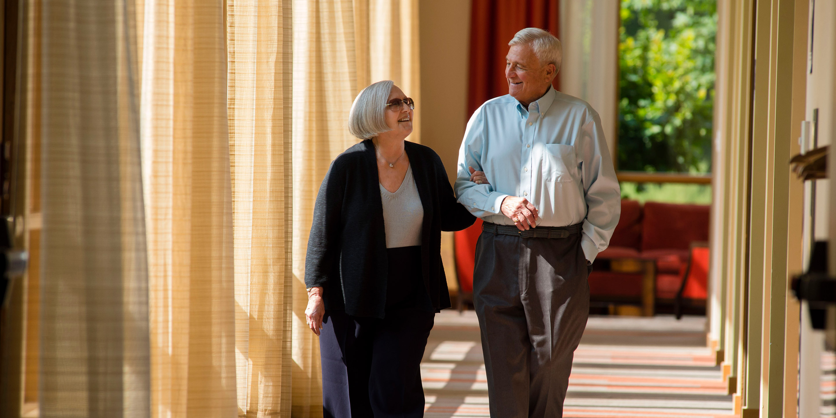 An older woman links her arm through an older man's arm as they walk down a hallway smiling.