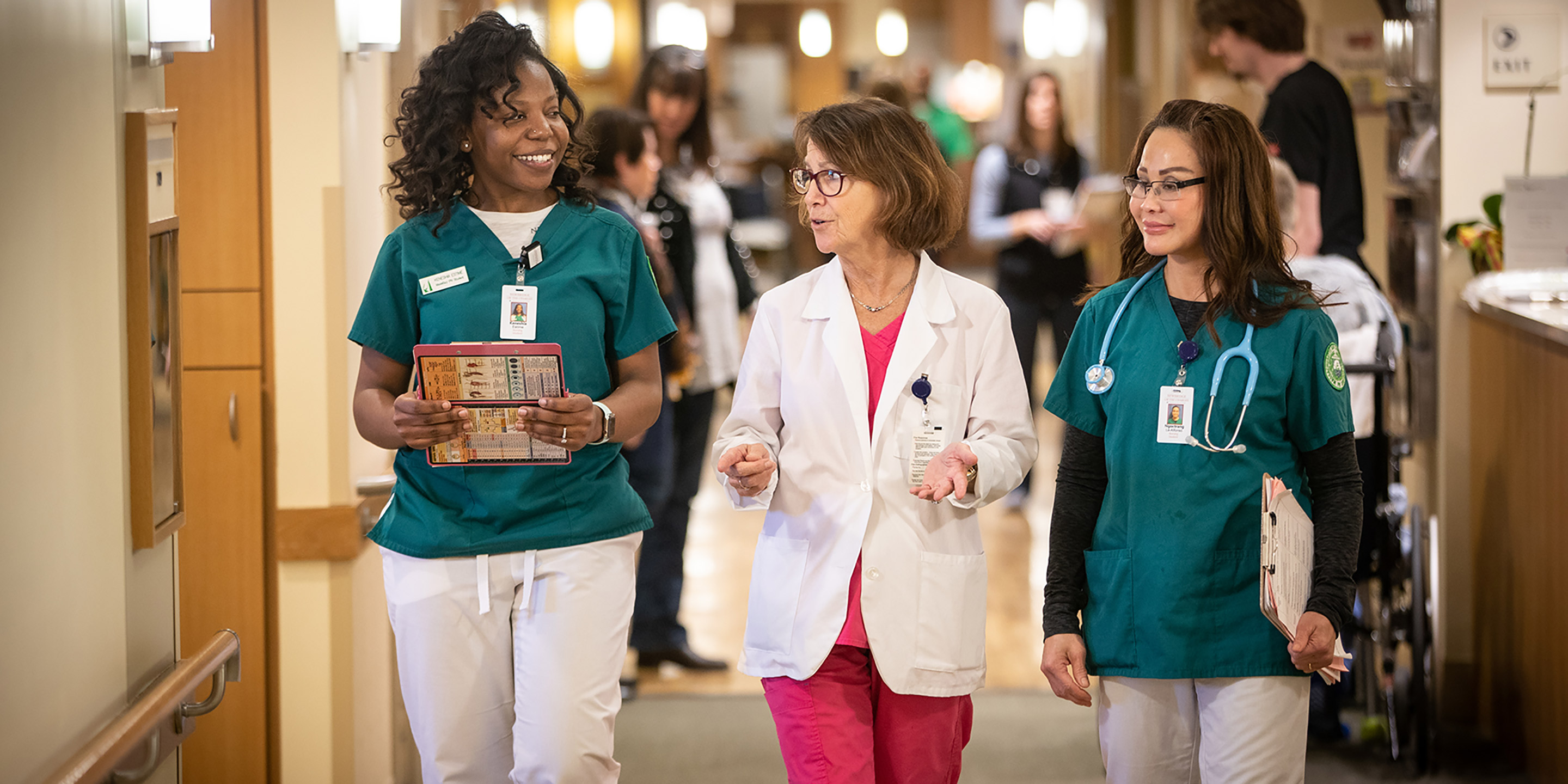 Three female nurses wearing scrubs walk down a hallway together.