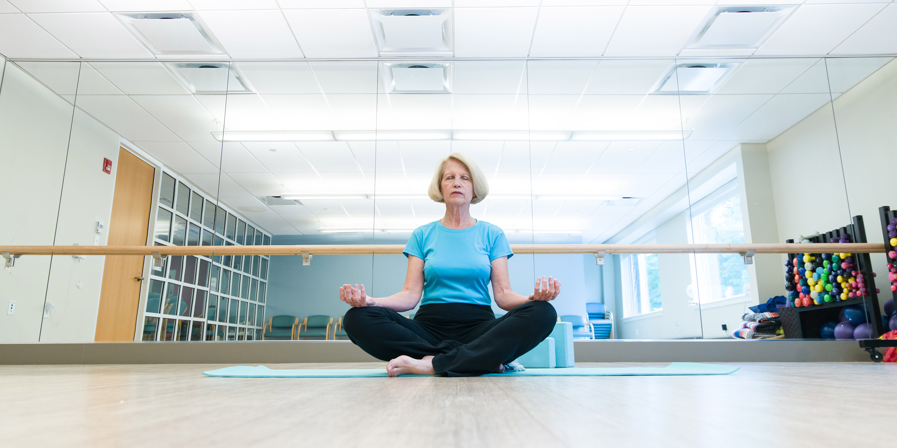 An older woman sits cross-legged and meditating on the floor of a gym at Orchard Cove in Canton, with her eyes closed and hands on her knees.