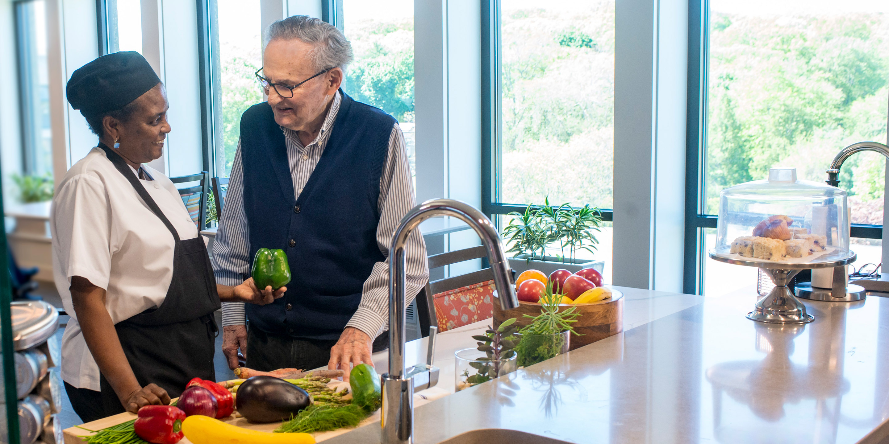 A male patient stands in the kitchen of his household at Hebrew Rehabilitation Center in Boston. A female member of the culinary team stands next to him, holding a green pepper. In front of them there are a variety of colorful fruits and vegetables on the counter. They are looking at each other and smiling.