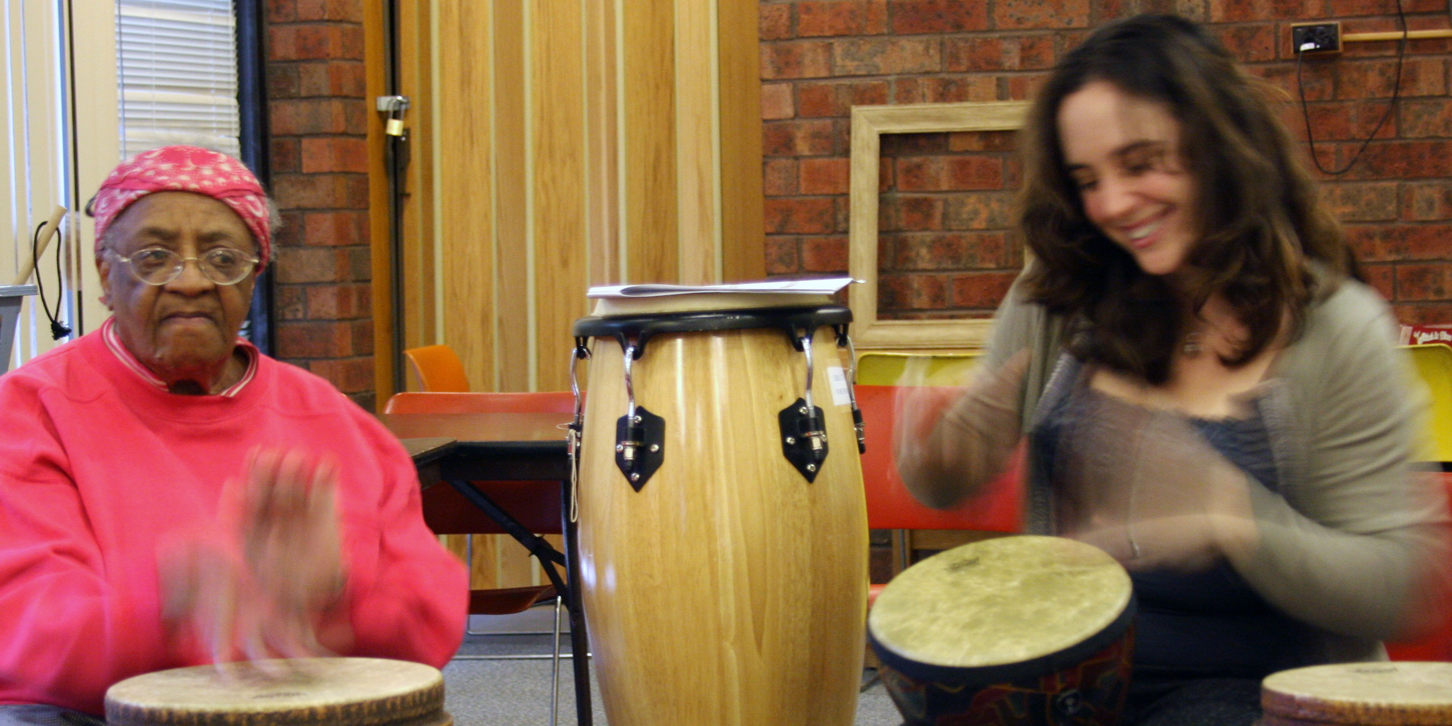 An older woman and a music therapist smile as they play the drums.