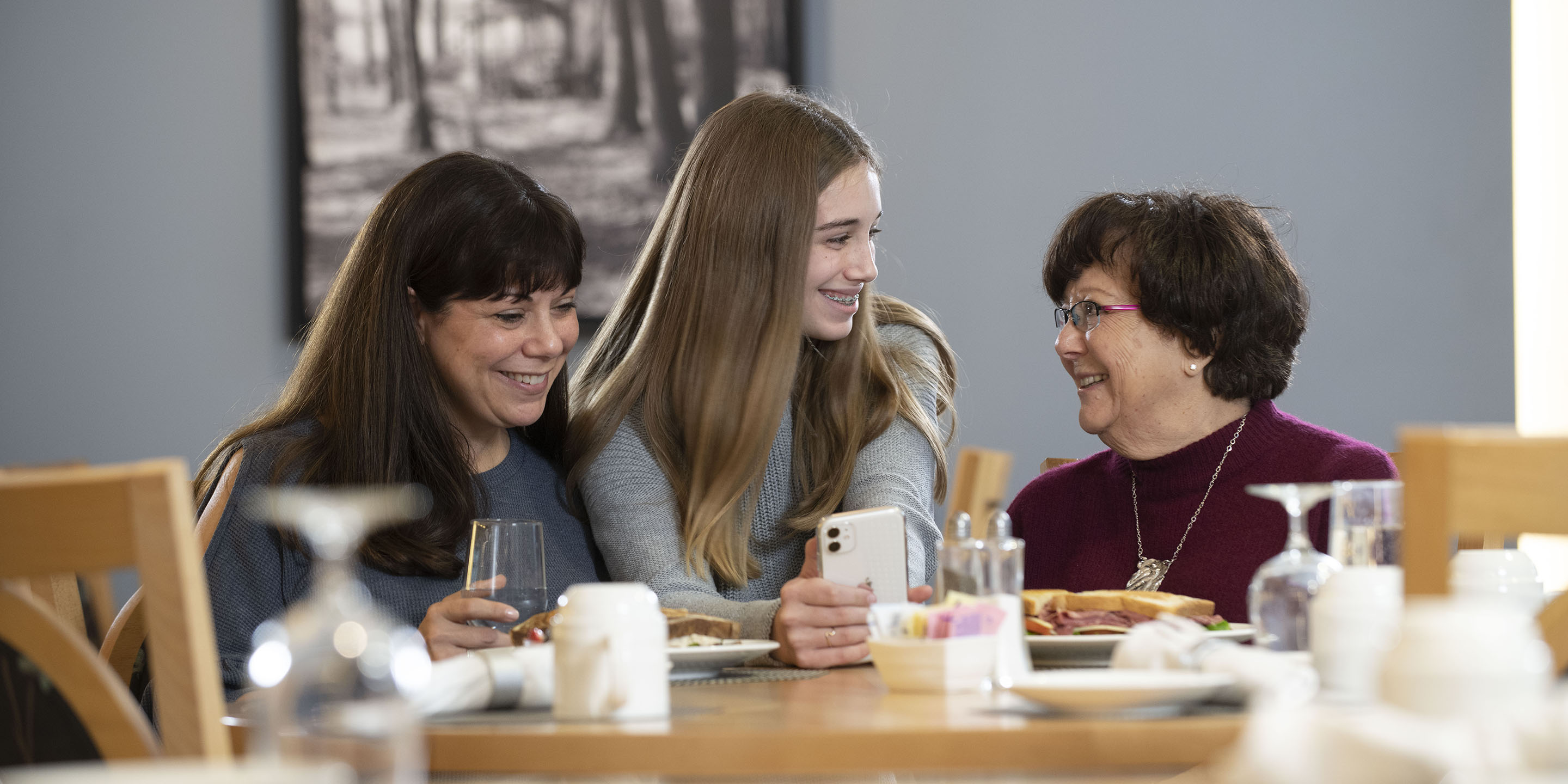 Grandmother, mother, and daughter sitting at a dinner table and looking at the daughter's cell phone. 