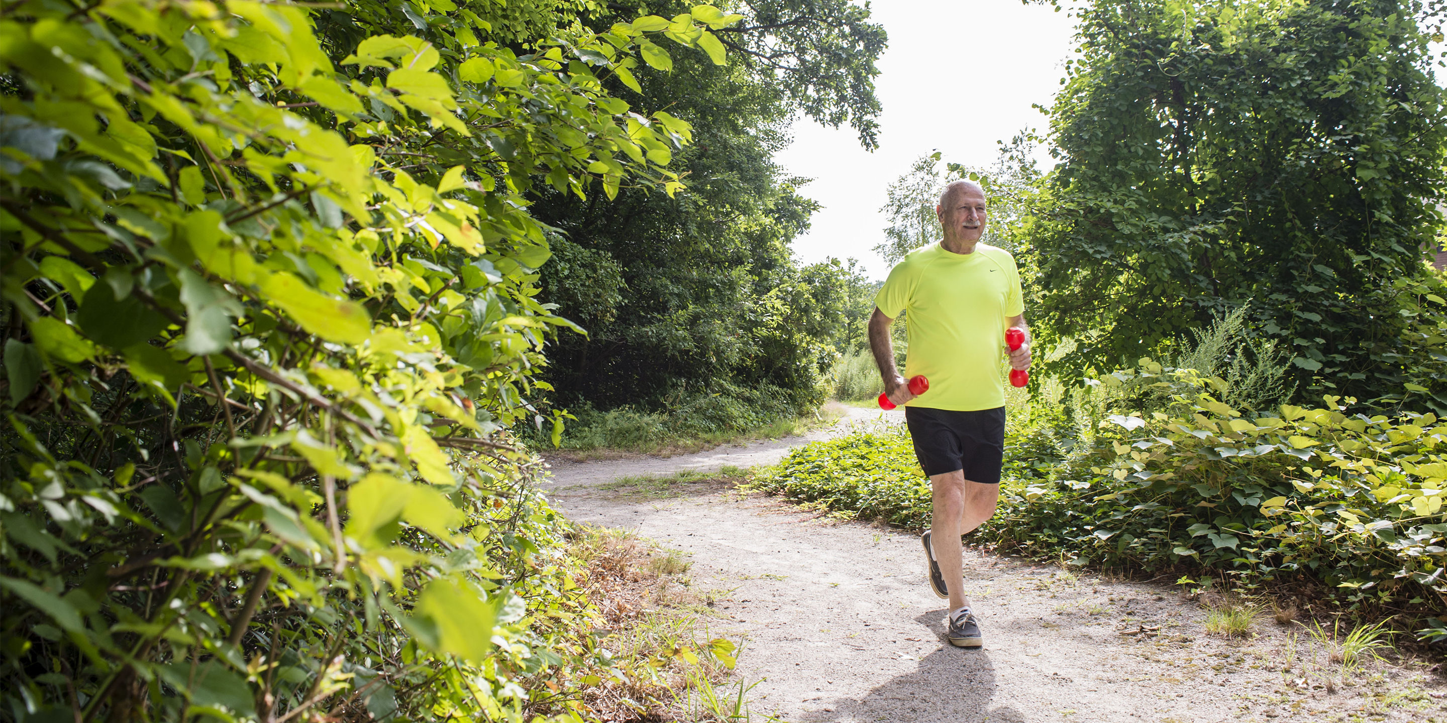 Older man with weights running on gravel path outside with trees.