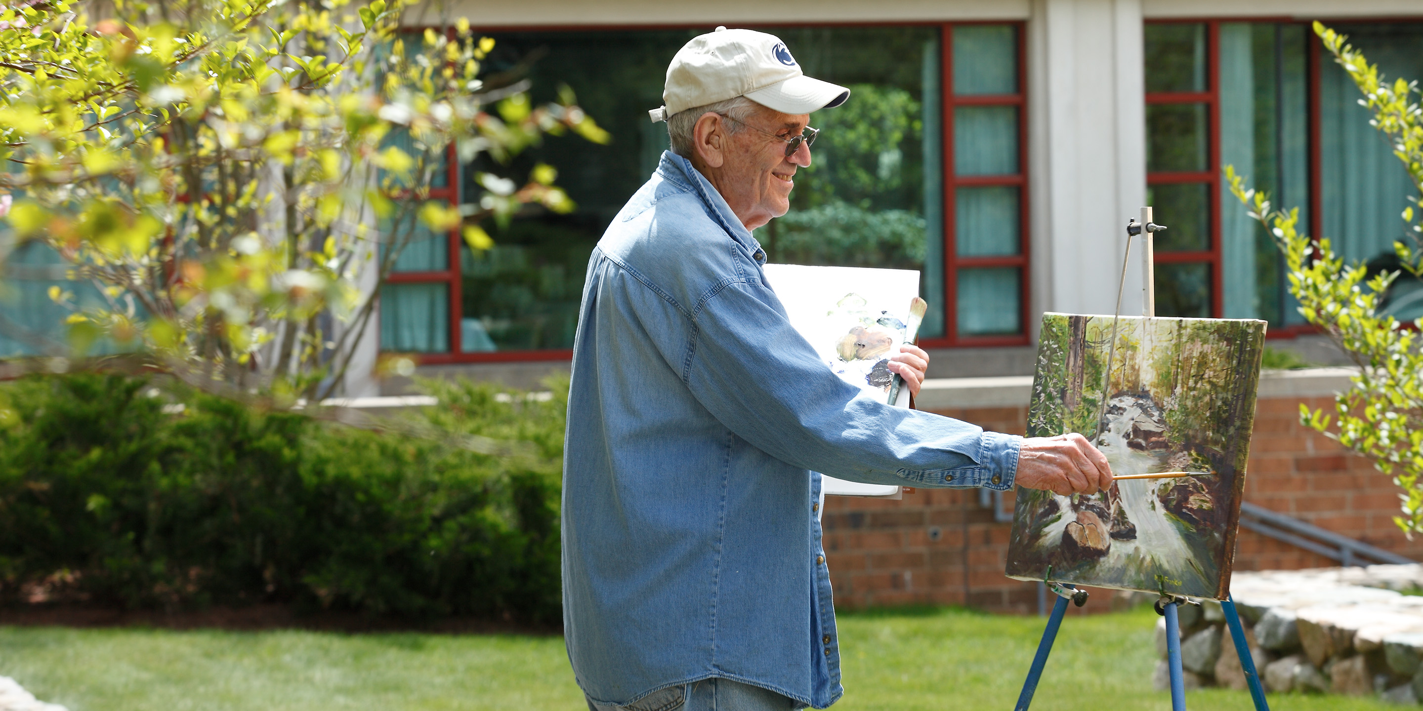 Man wearing blue clothing and a ballcap holding a painter’s palette in the process of painting a waterfall while standing by a tree outside his apartment.