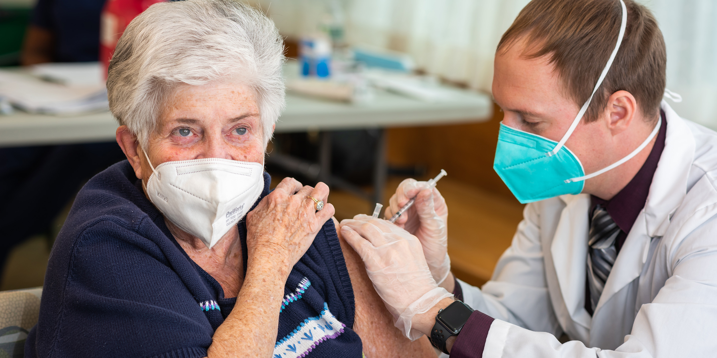 Woman in a blue shirt wearing a mask receives COVID-19 vaccine administered by man with a tie and labcoat.