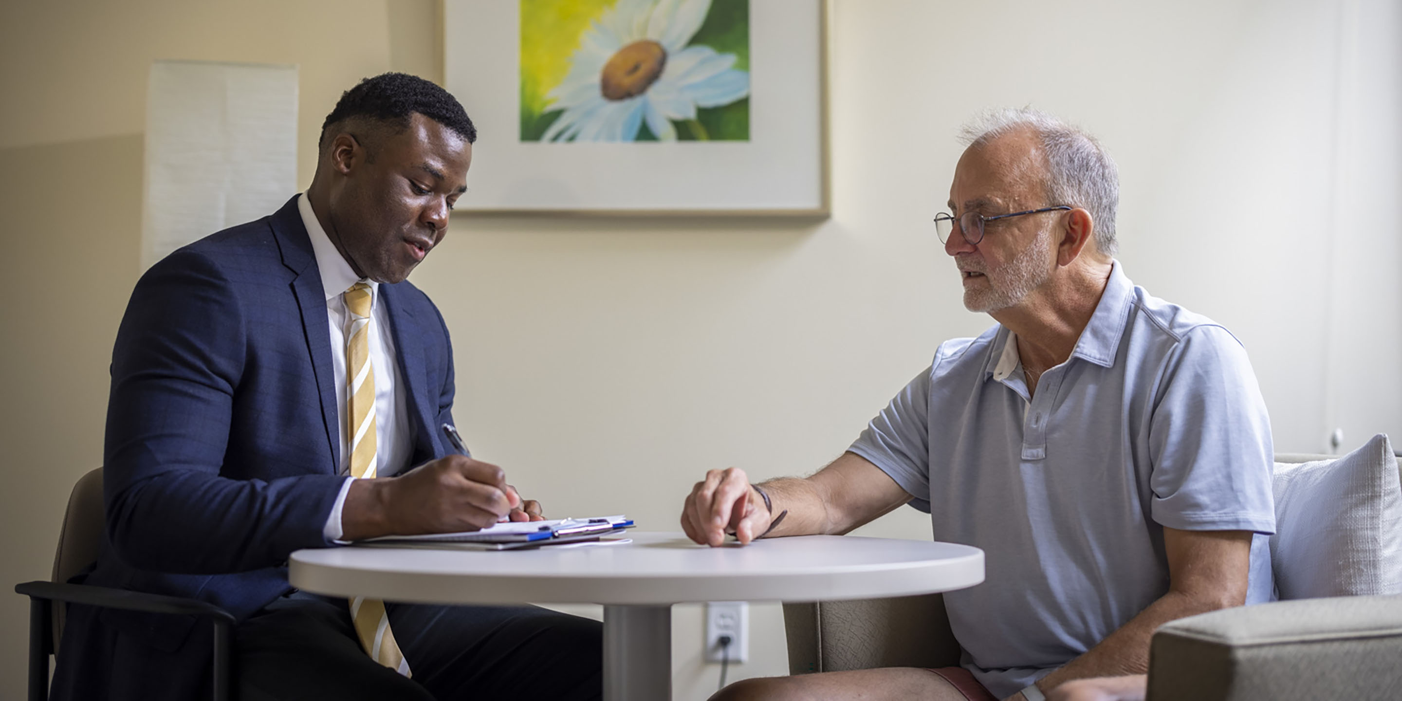 Older man sitting at table with doctor in blue blazer discussing report on clipboard. 