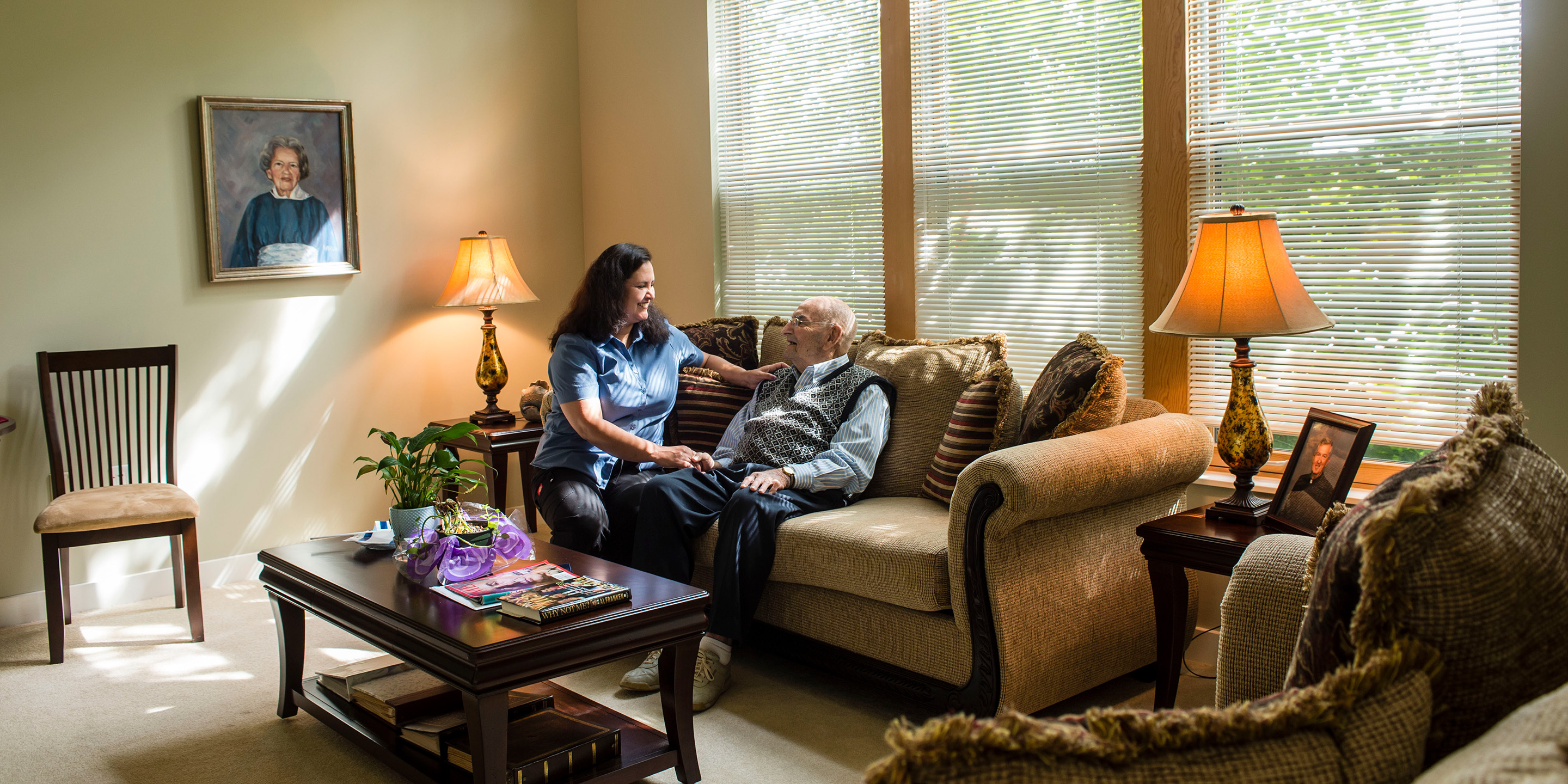 Woman with a hand on the shoulder and holding the hand of an older man sitting on a couch in a living room