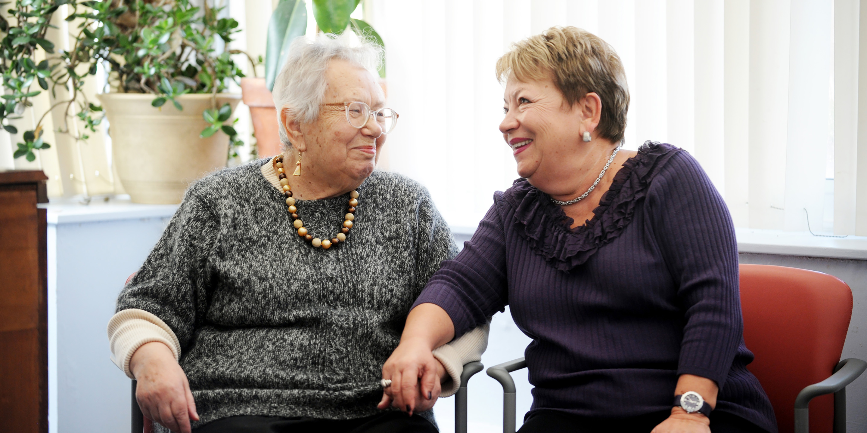 Two women sitting in chairs holding hands, smiling and talking to each other.