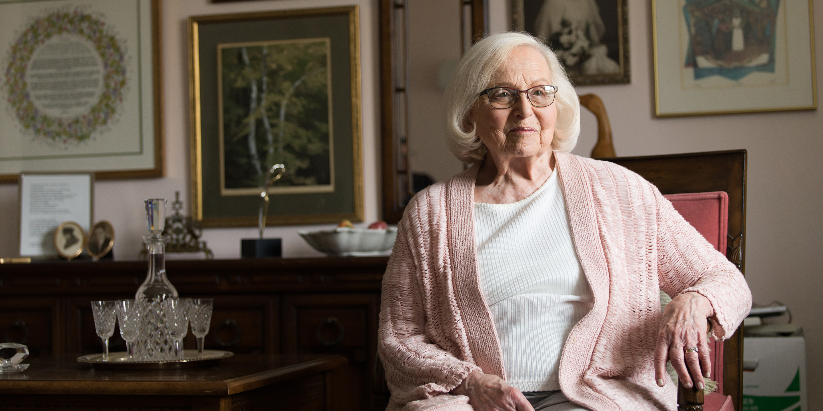 Smiling woman wearing pink sweater sitting in a chair next to a crystal decanter and goblets with framed pictures of landscapes and people on the wall behind her.